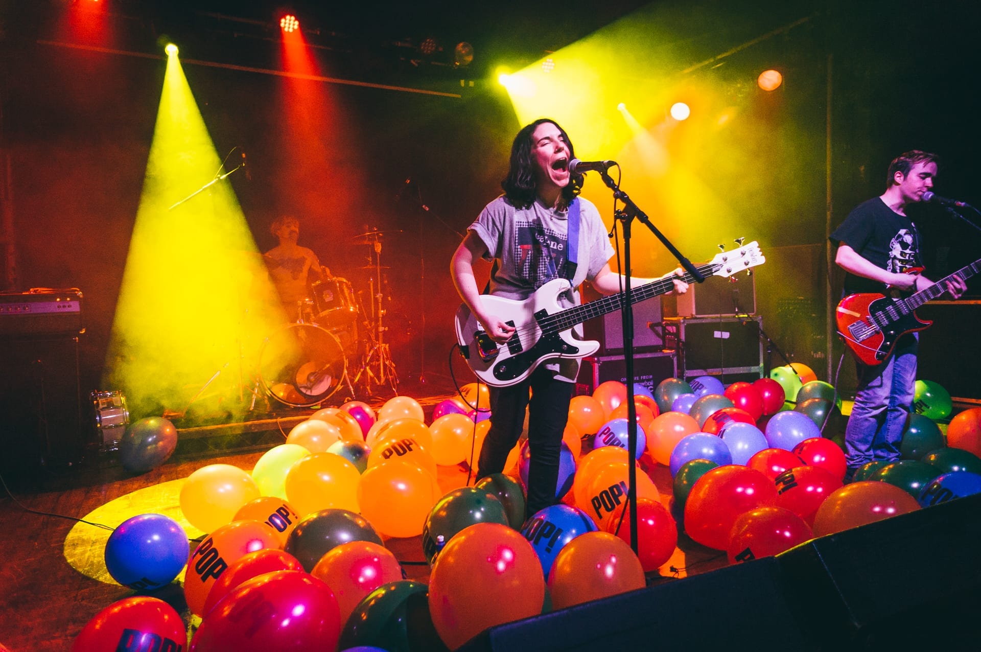 The Spook School play a stage covered in balloons printed with the word POP, a bass player singing at the centre mic, a guitarist at right and a drummer behind.