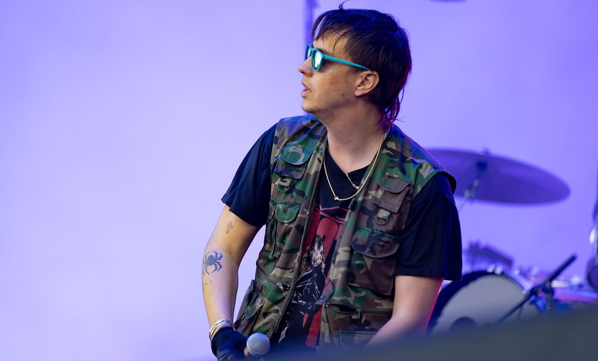 A member of The Strokes performing live in profile, wearing tinted sunglasses and a camouflage vest over a black T-shirt, holding a microphone, with a drum kit visible behind him under purple stage lighting.
