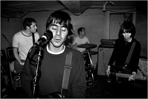 The Van Pelt perform live in a small basement venue, in a black and white photograph: the singer plays guitar and sings into a microphone in the foreground, with a drummer and bassist behind him.