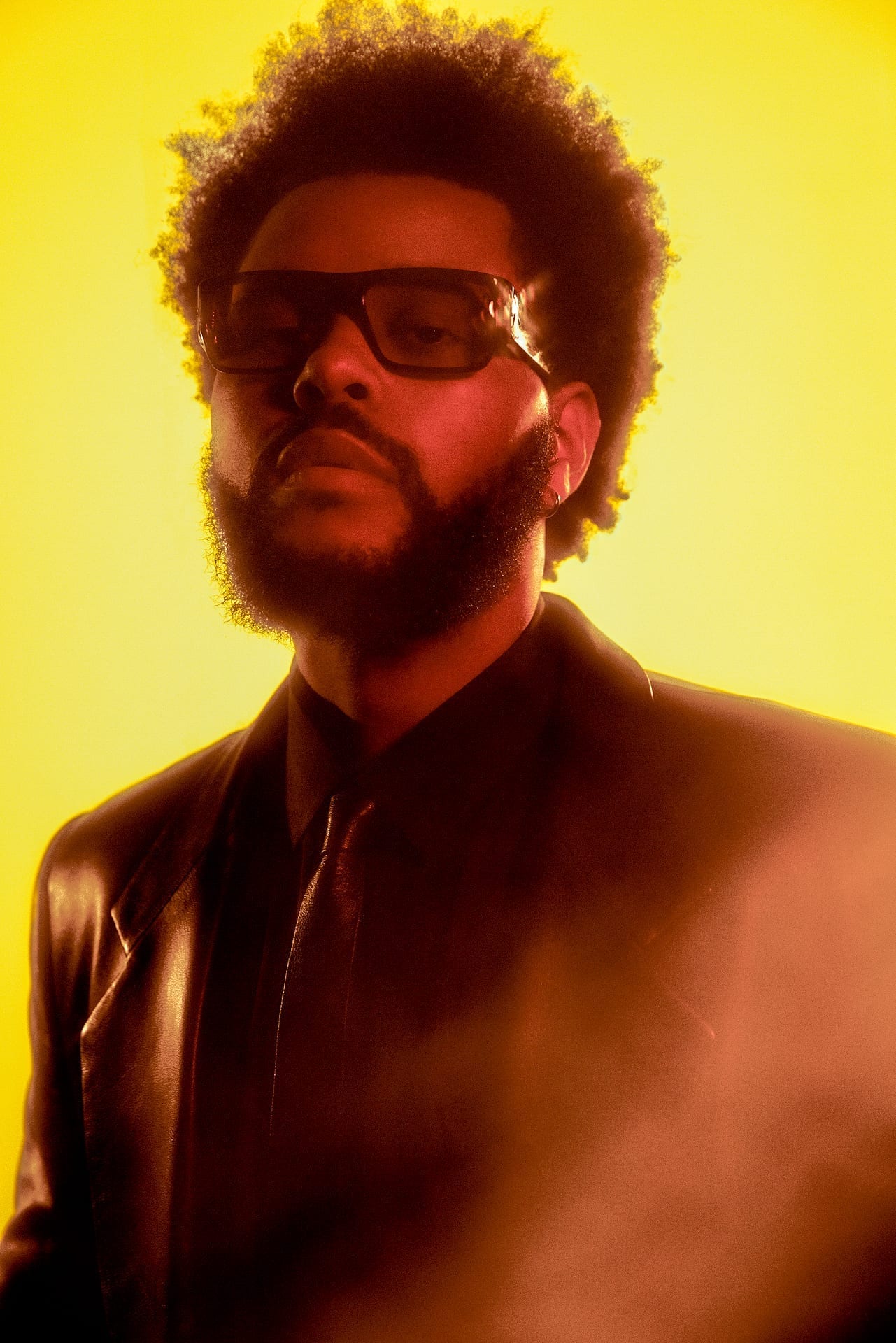 The Weeknd, wearing dark glasses and a black leather jacket, looks into the camera in a moody portrait lit in yellow and red.