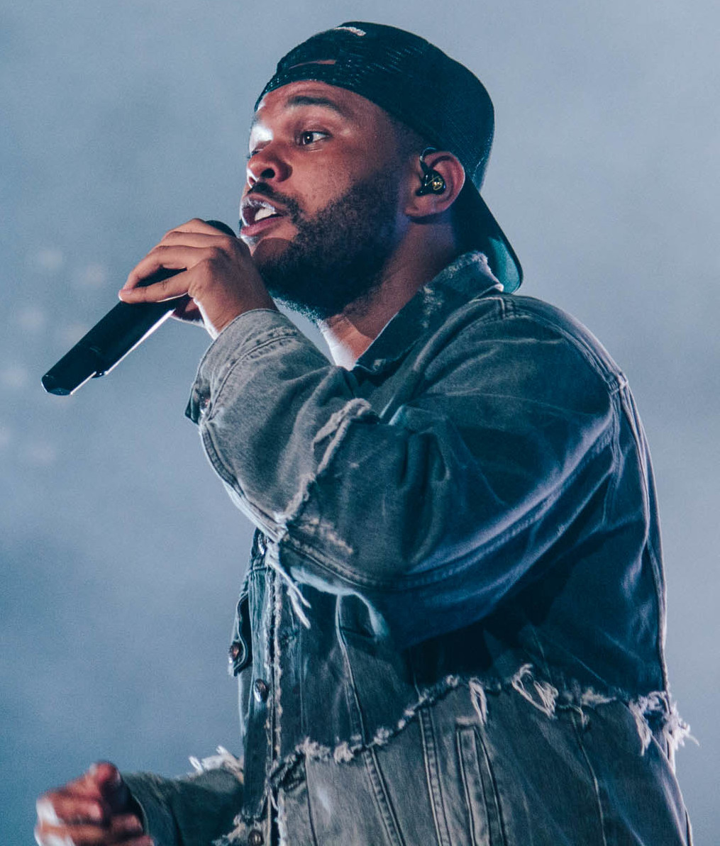 A singer in a distressed denim jacket and a backwards cap sings into a handheld microphone, lit against a pale hazy stage background