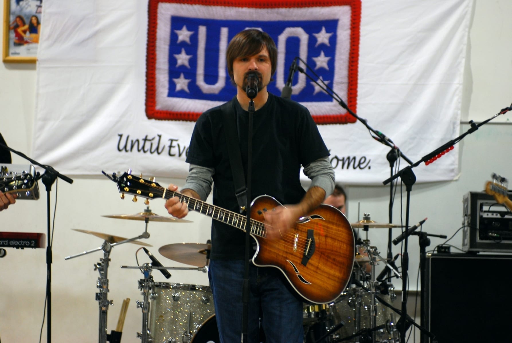 Third Day frontman Mac Powell sings into a microphone while playing an acoustic-electric guitar on stage, with a drum kit visible behind him.
