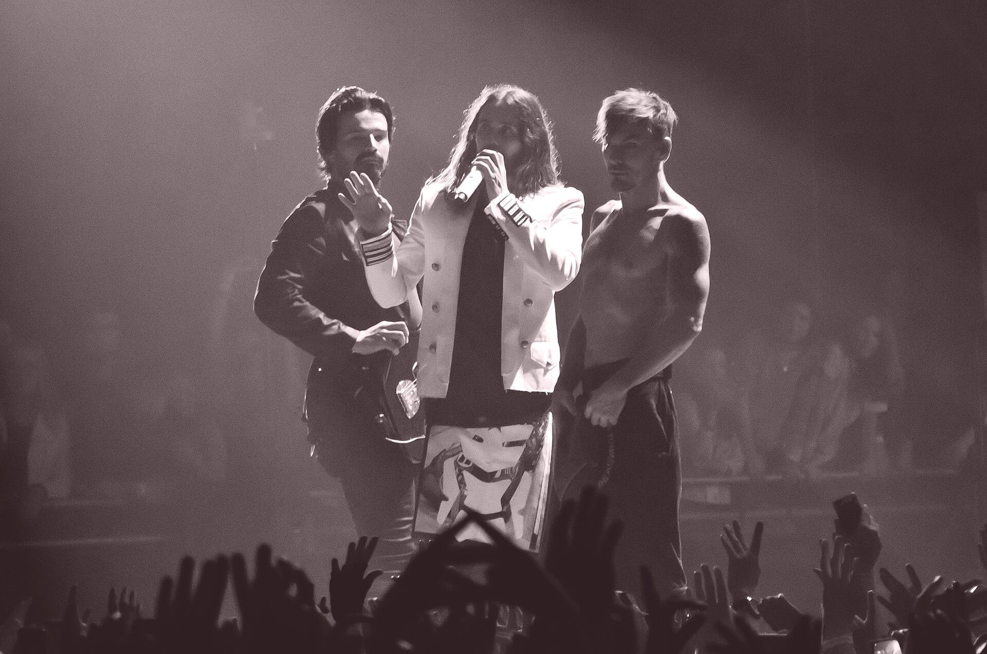 Thirty Seconds to Mars perform on stage in black and white: one member holds a guitar, the singer holds a microphone with a hand raised, and a shirtless member stands to the side, with a crowd's raised hands visible in the foreground.