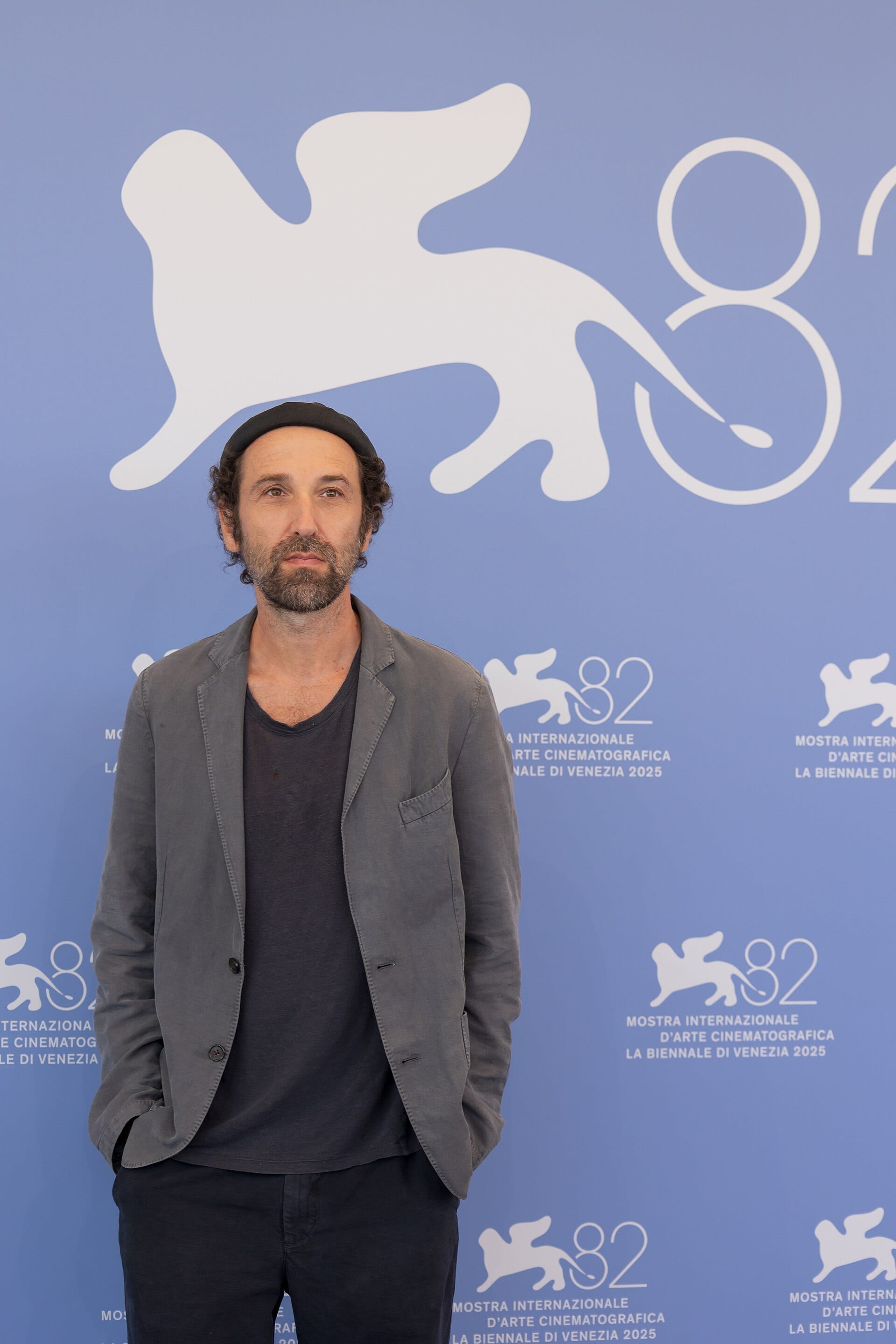 Thomas Bangalter, in a gray blazer and dark cap, stands for photos in front of a blue backdrop bearing the Venice Film Festival's winged-lion logo.