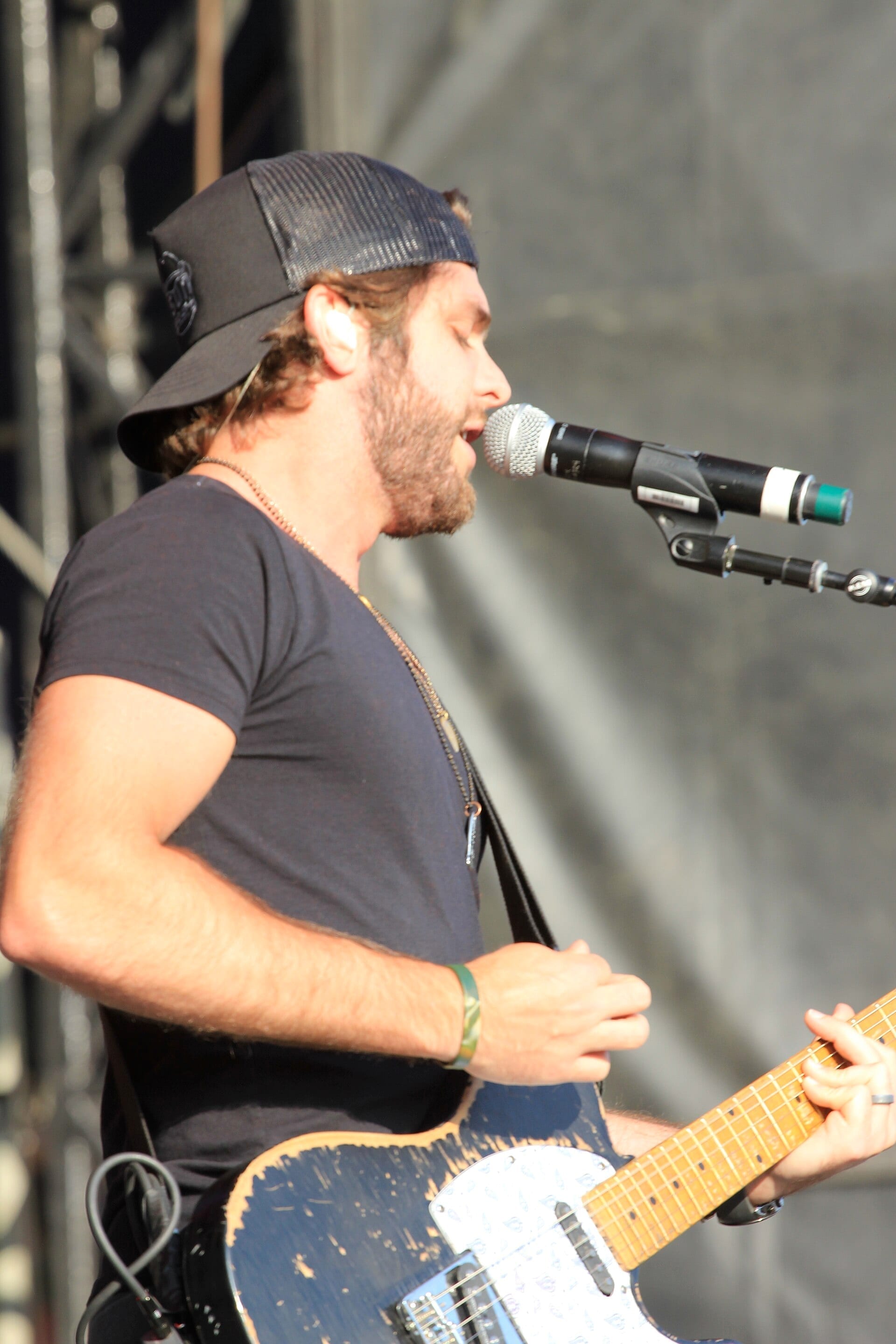 Thomas Rhett, wearing a backwards cap and a dark t-shirt, sings into a microphone in profile while playing a battered blue electric guitar on an outdoor stage.