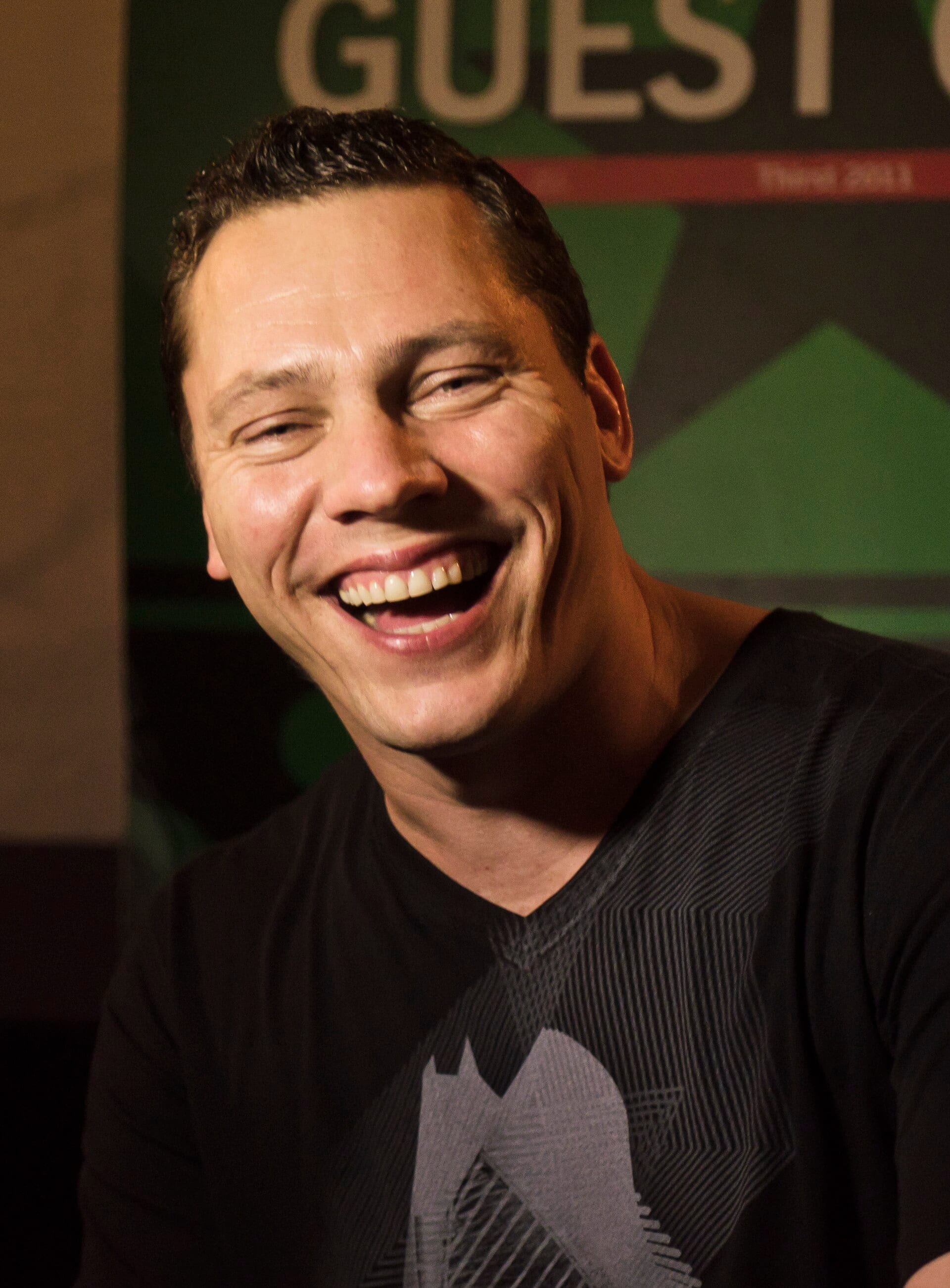 Tiesto laughs while wearing a black V-neck T-shirt, standing in front of a green backstage sign reading Guest.