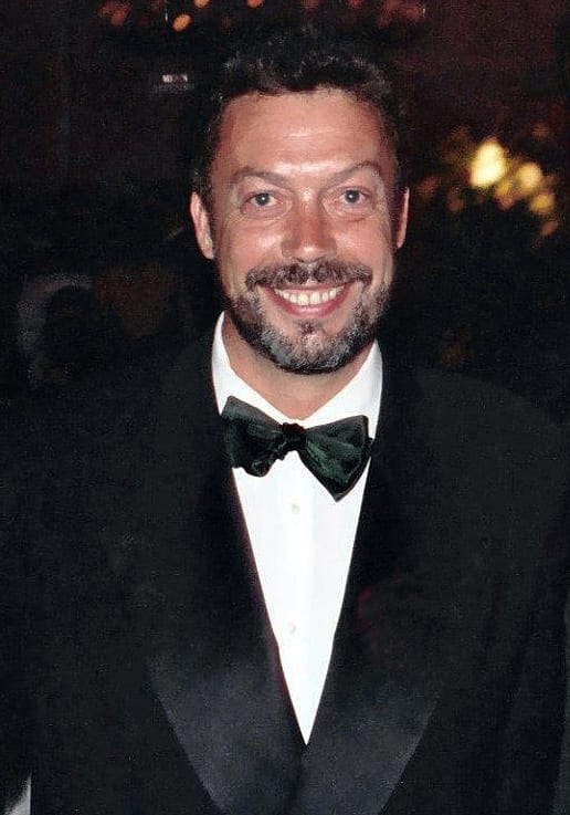 Tim Curry smiles at the camera in a black dinner jacket and a dark green velvet bow tie, a lit room out of focus behind him.