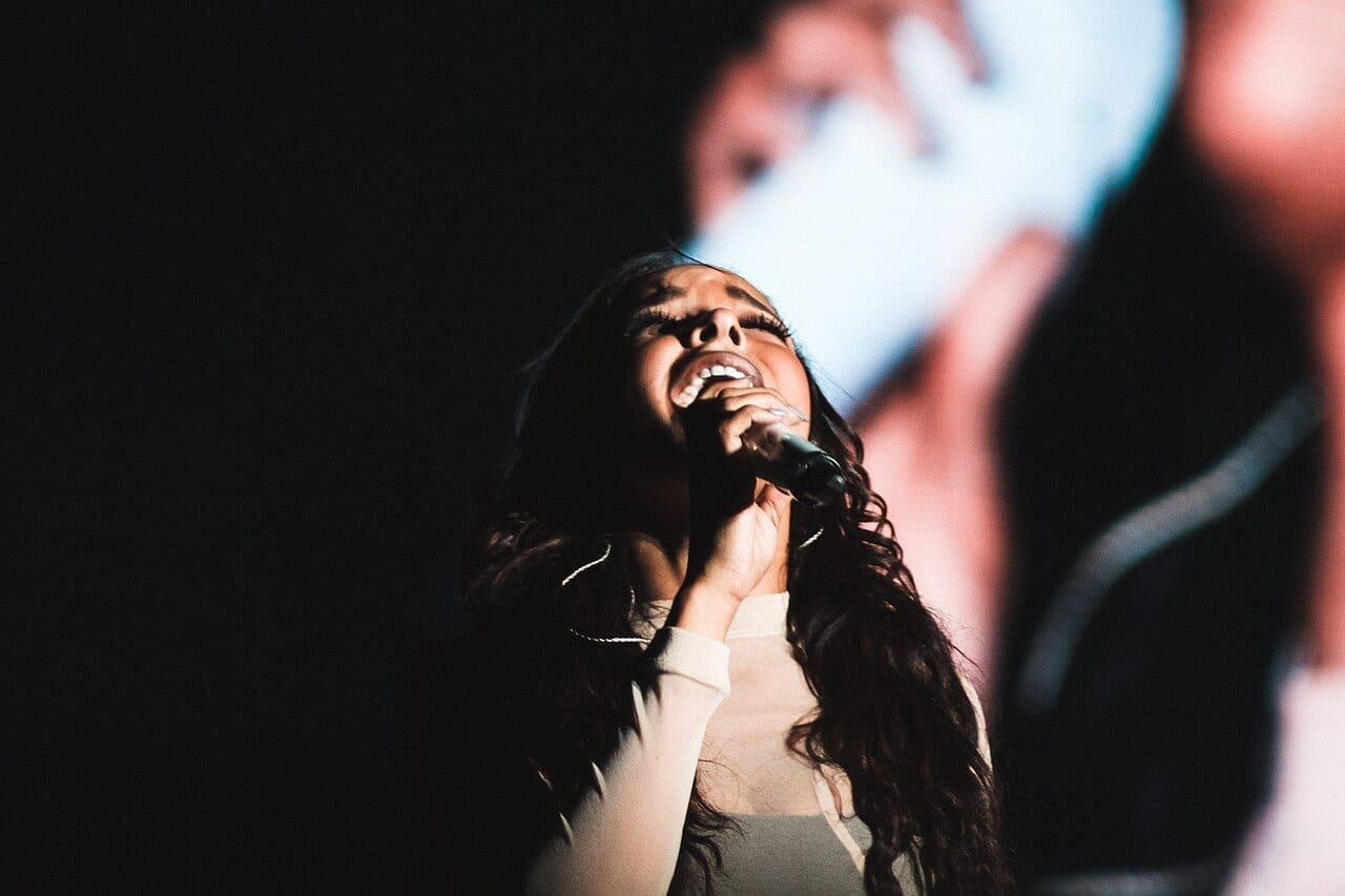 Tinashe sings into a handheld microphone with her head tilted back, lit against a dark stage.