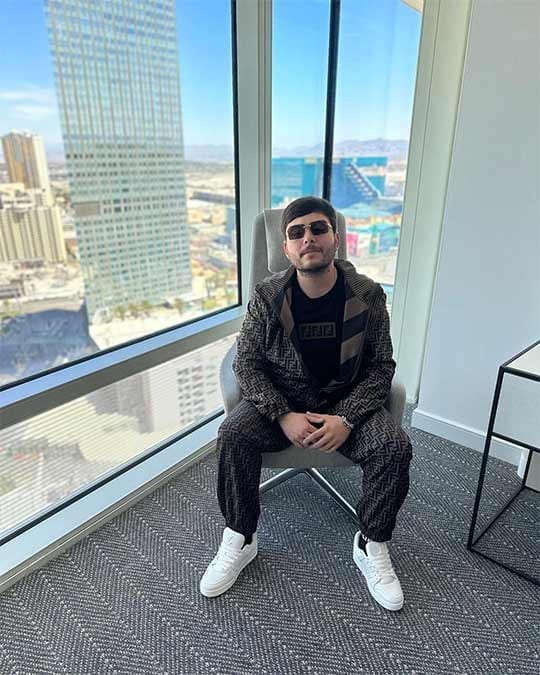 Tito Double P, wearing sunglasses and a matching patterned tracksuit, sits in a chair by a floor-to-ceiling window overlooking a city skyline.