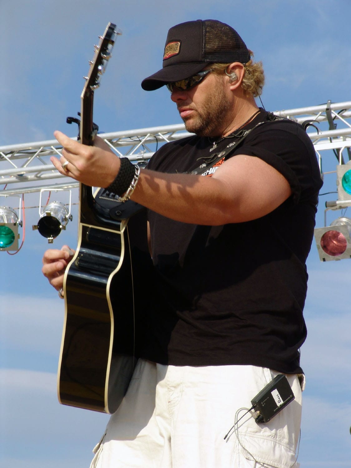 Toby Keith plays a black acoustic guitar on an outdoor stage, wearing a dark trucker cap, sunglasses and a black sleeveless shirt, with stage lighting rigging visible behind him against a blue sky.