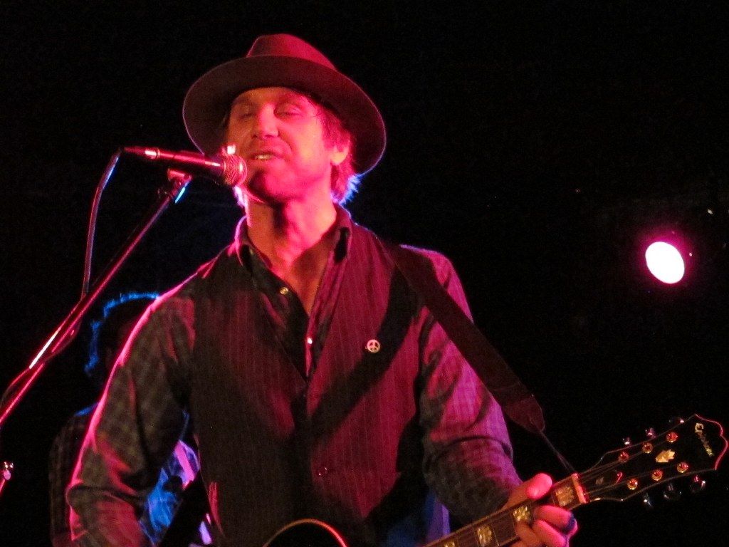 Todd Snider sings into a microphone in a wide-brimmed hat and a striped shirt, a guitar neck in his hand under red stage light.