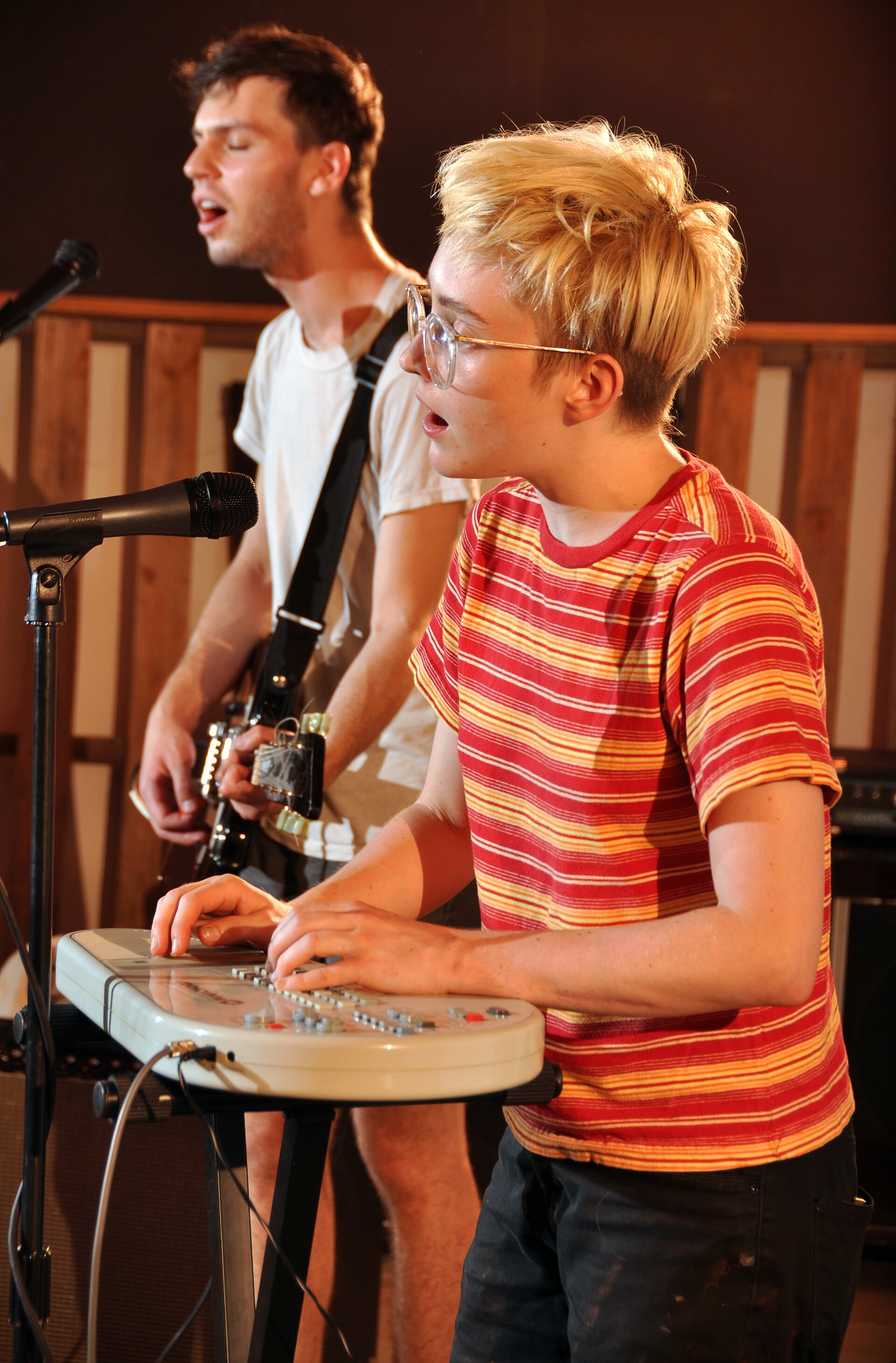 Two members of Told Slant play a small room, one singing over a keyboard and the other beside them, lit warm against a dark wall.
