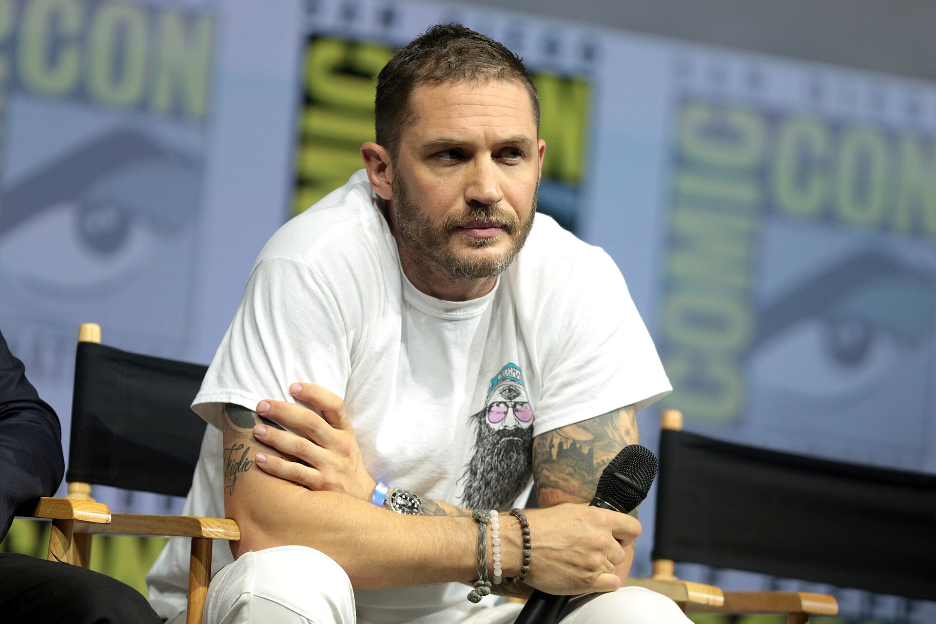 Tom Hardy sits in a director's chair with his arms folded and a microphone in one hand, in a white printed T-shirt, a Comic-Con backdrop behind him.