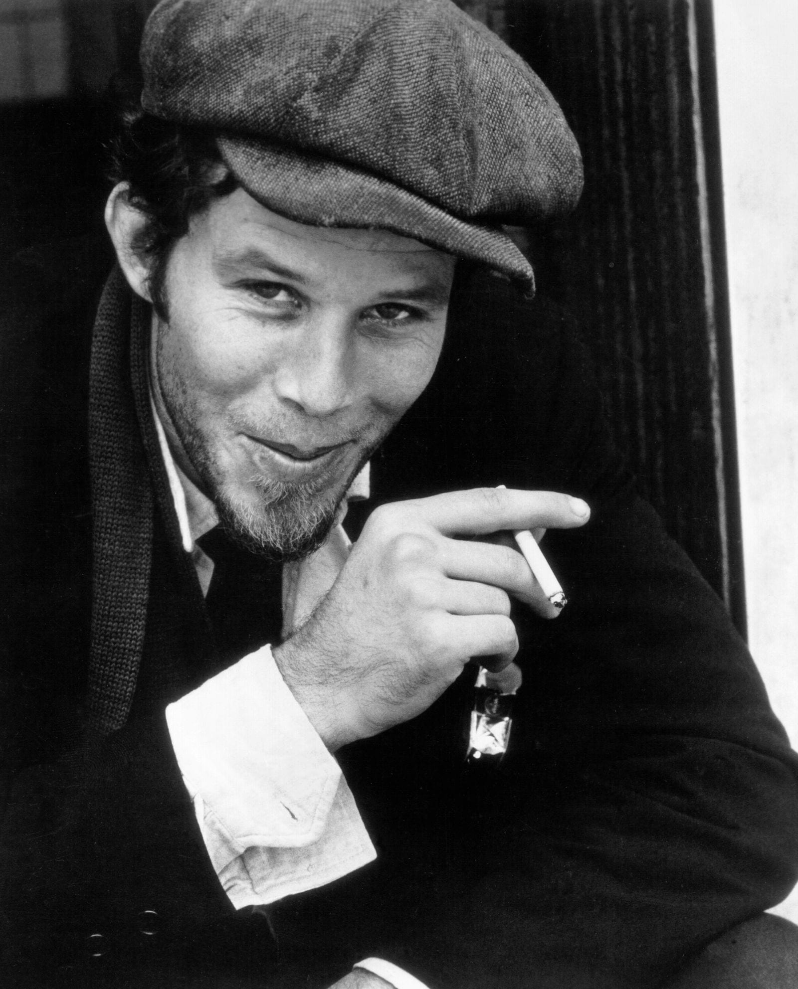 A young Tom Waits, wearing a flat cap and a scarf, smiles slightly while holding a lit cigarette in a black-and-white portrait.