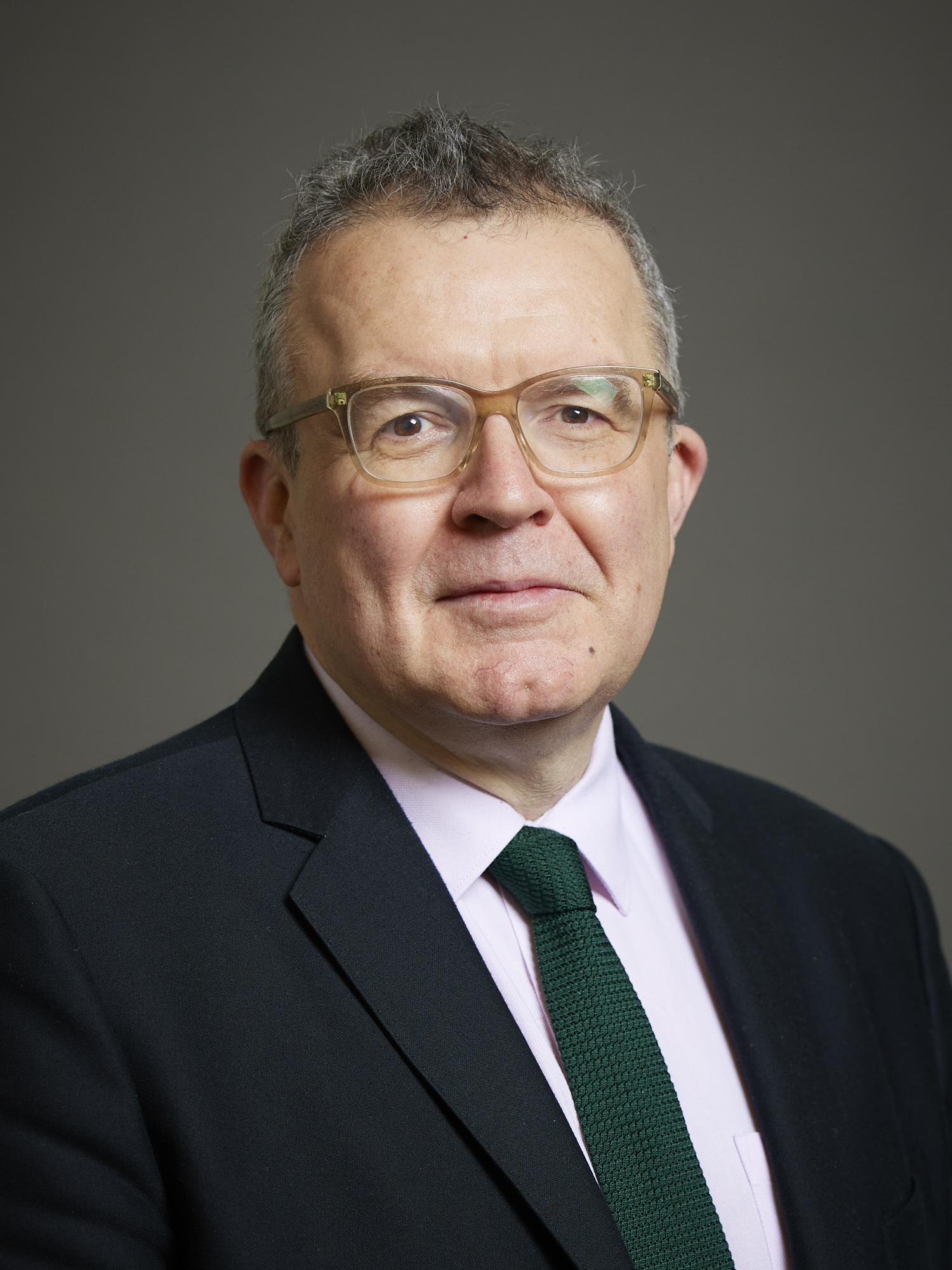 A grey-haired man in clear-framed glasses, a dark suit and a green knitted tie looks at the camera against a plain grey studio background