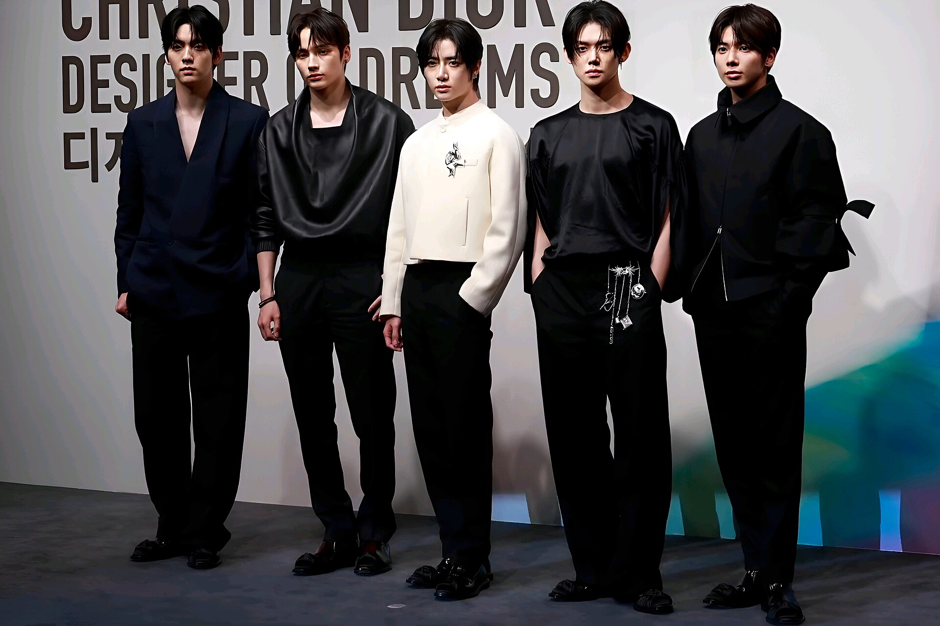 The five members of Tomorrow X Together stand in a line in black and cream outfits in front of a Christian Dior backdrop.