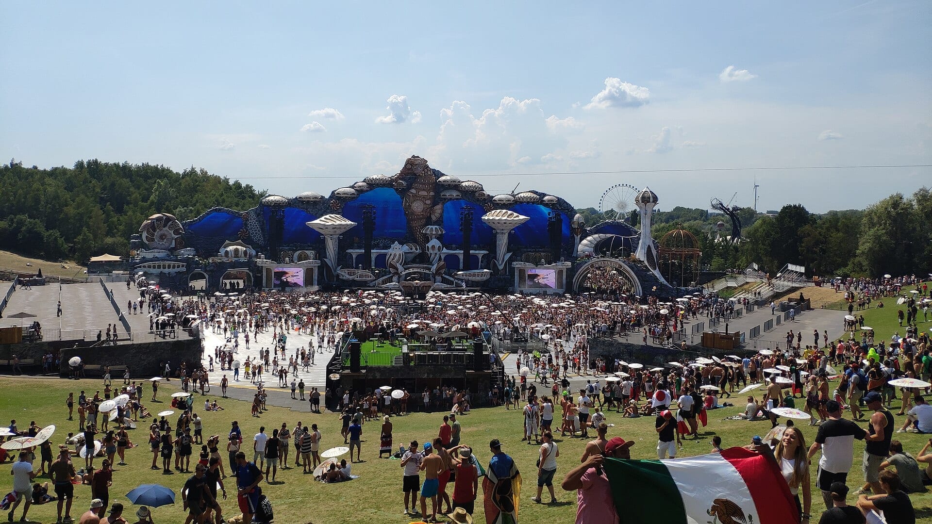 Tomorrowland's main stage, a wide sculpted set of shells and towers, faces a crowd spread across grass and paving under a bright sky, with a ferris wheel behind the treeline.