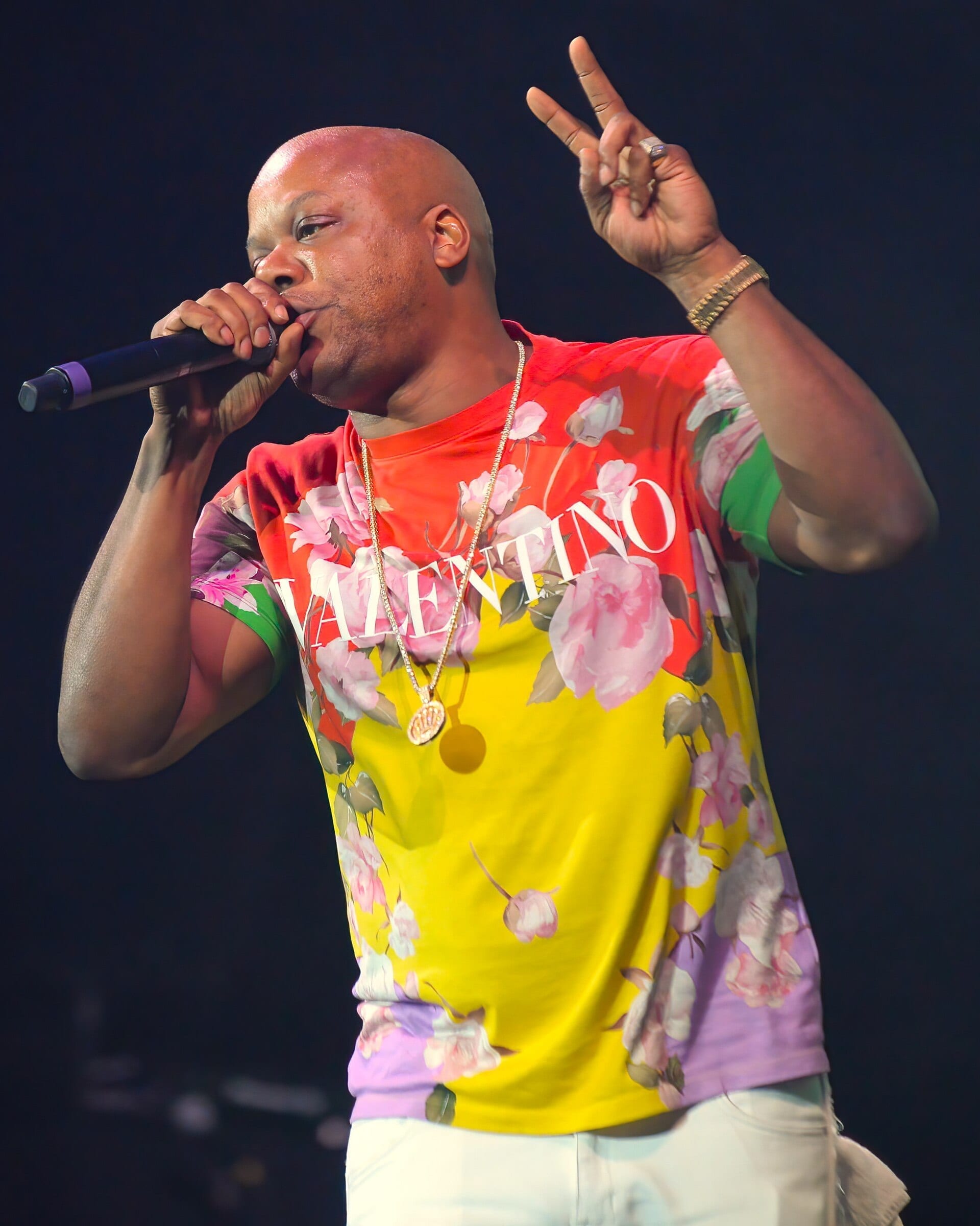 Too Short raises a hand in a peace sign while singing into a microphone on stage, wearing a brightly colored floral T-shirt.