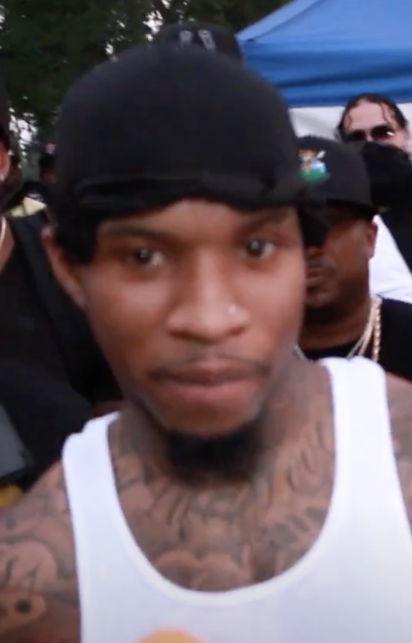 A rapper in a white vest and a backwards black cap looks straight at the camera outdoors, chest and neck tattoos visible, people and a gazebo behind him