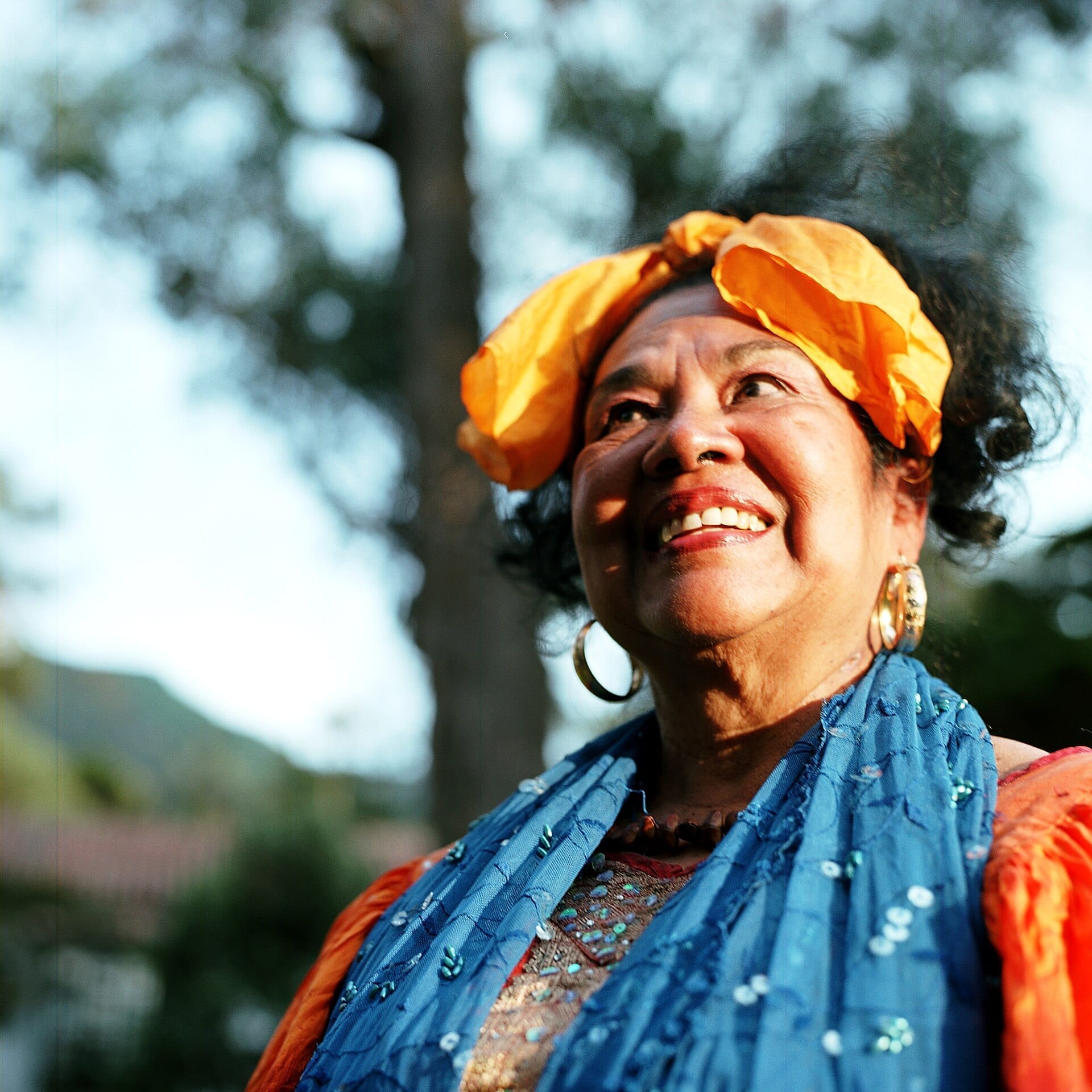 Toto La Momposina smiles and looks upward outdoors, wearing a bright orange headscarf, hoop earrings and a sequined blue shawl.