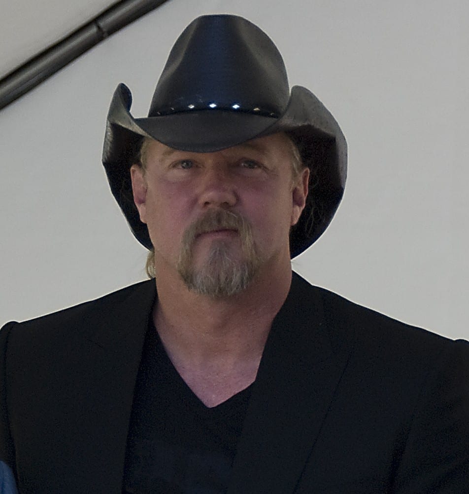 Trace Adkins looks straight ahead under a black cowboy hat, wearing a black blazer over a black V-neck, his grey goatee catching the light.