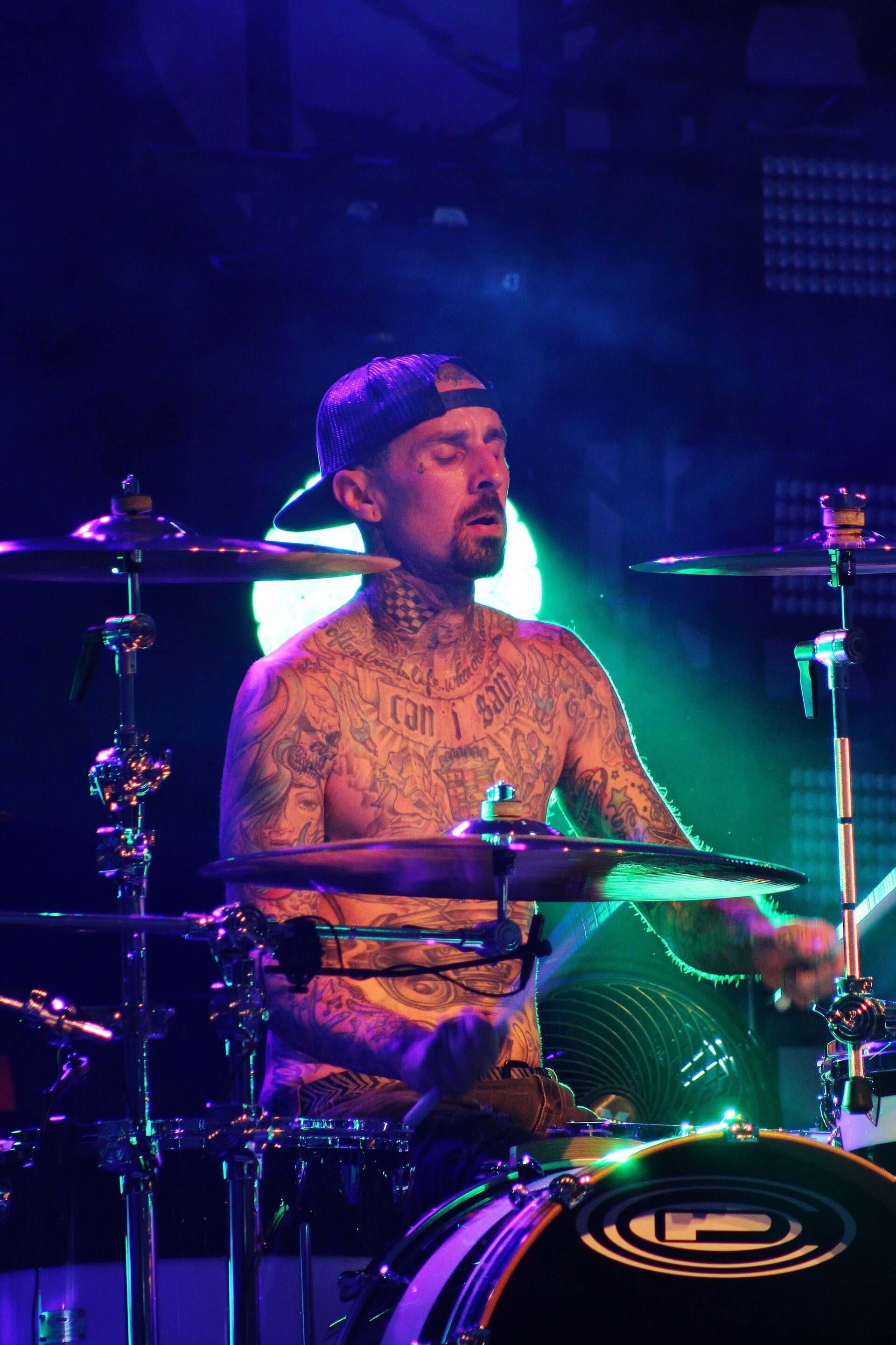 A heavily tattooed, shirtless Travis Barker plays drums on stage under blue and green lights, wearing a backwards cap.