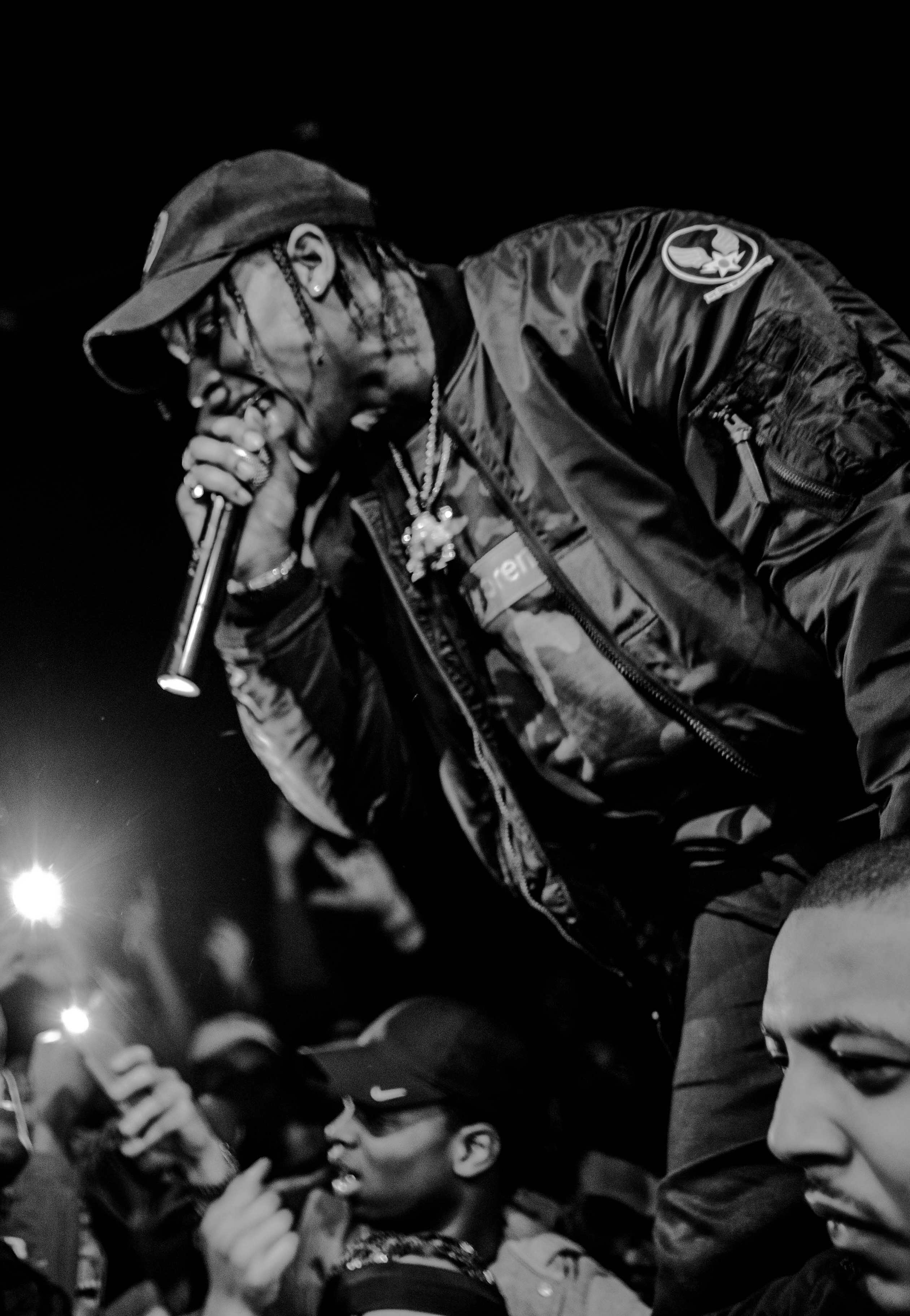 Travis Scott leans out over a crowd in a dark cap and bomber jacket, rapping into a microphone, in a black and white photograph.
