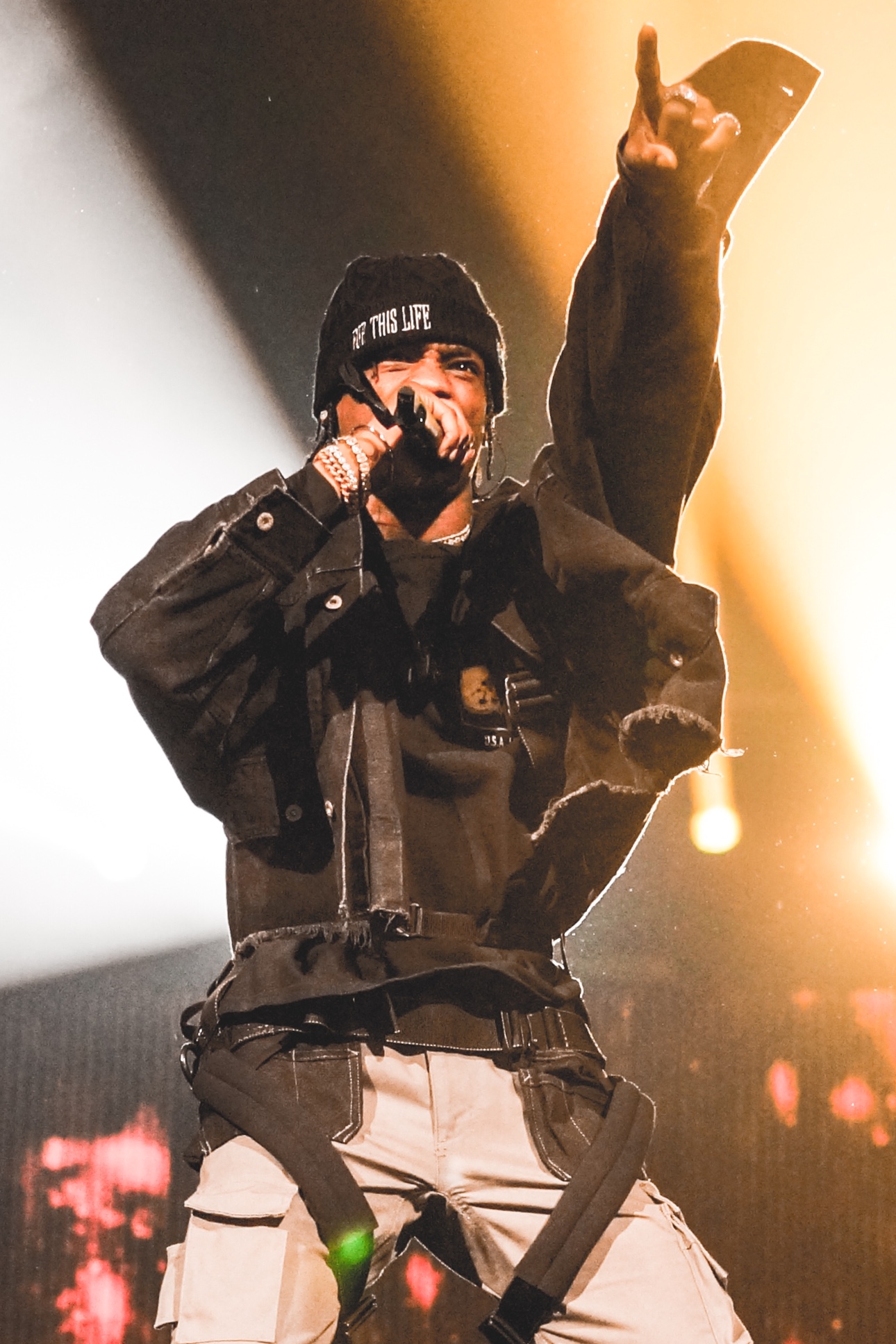 Travis Scott raps into a microphone with one arm thrown up, in a beanie and a black jacket over a climbing harness, gold light behind him.