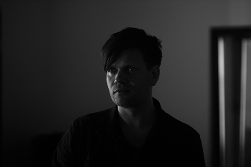 A dark, low-contrast portrait of Anders Trentemoller looking straight at the camera in a dim room.
