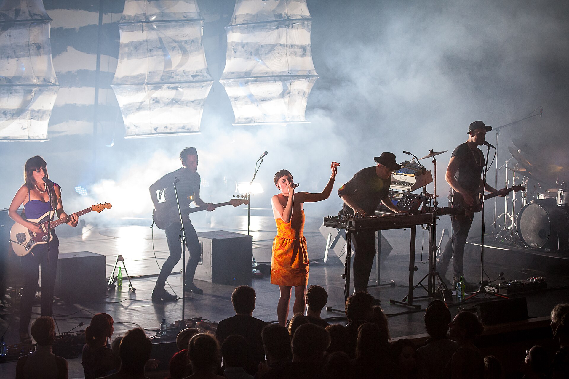 A five-piece band plays a concert hall stage in low blue light, a singer in an orange dress at the front microphone and a player in a wide-brimmed hat behind the keyboards.