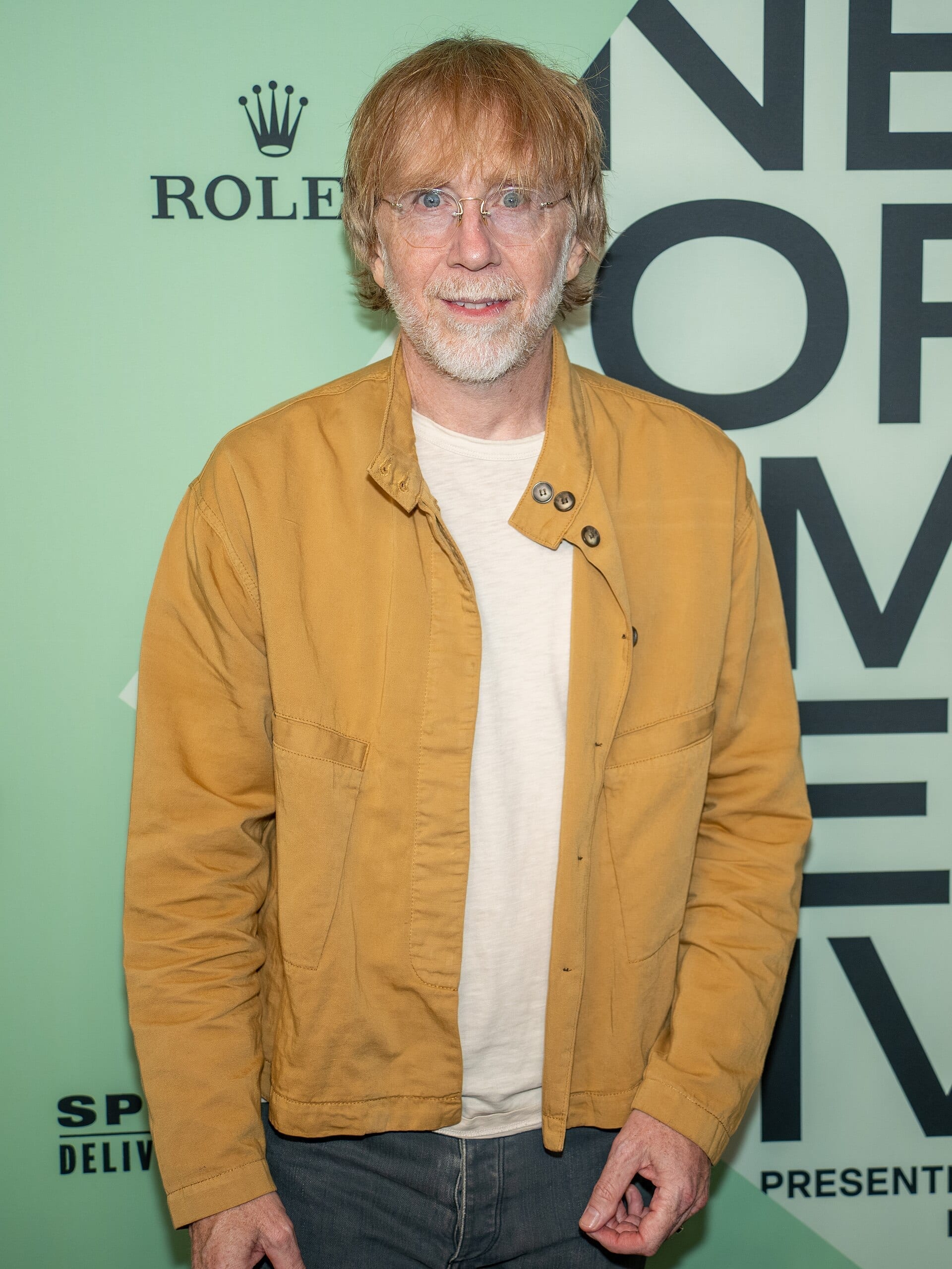 Trey Anastasio, with tousled reddish hair, wire-rim glasses and a gray beard, smiles at the camera in a mustard-colored jacket at a step-and-repeat event.