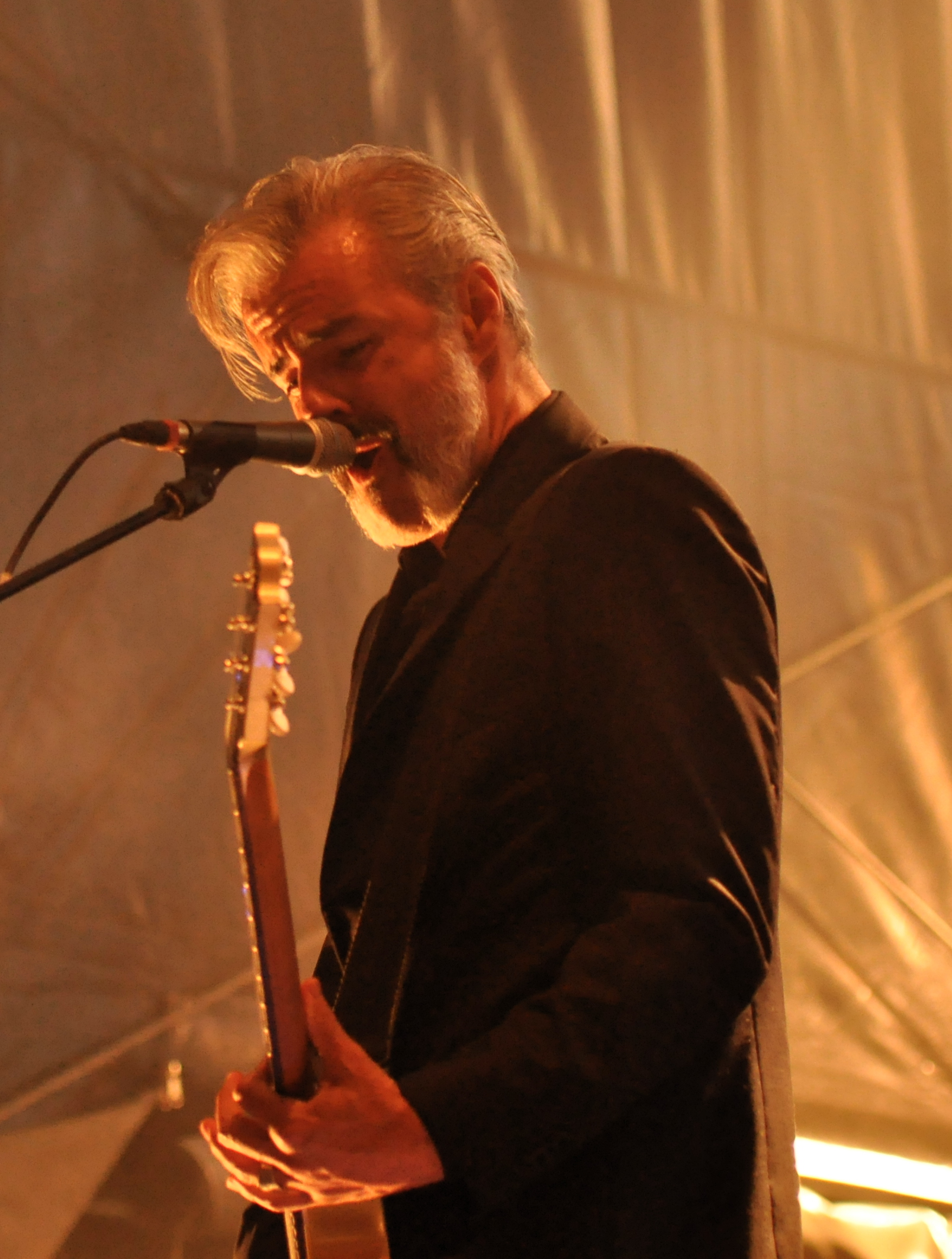 Ruben Block of Triggerfinger singing into a microphone under warm light in a black suit, grey hair swept back and a guitar neck held upright in front of him