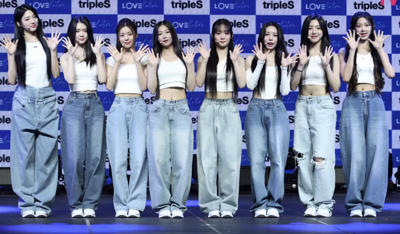 Eight members of tripleS standing in a row on a showcase stage in matching white tops and loose blue jeans, hands raised in a wave, a tripleS-branded backdrop behind them