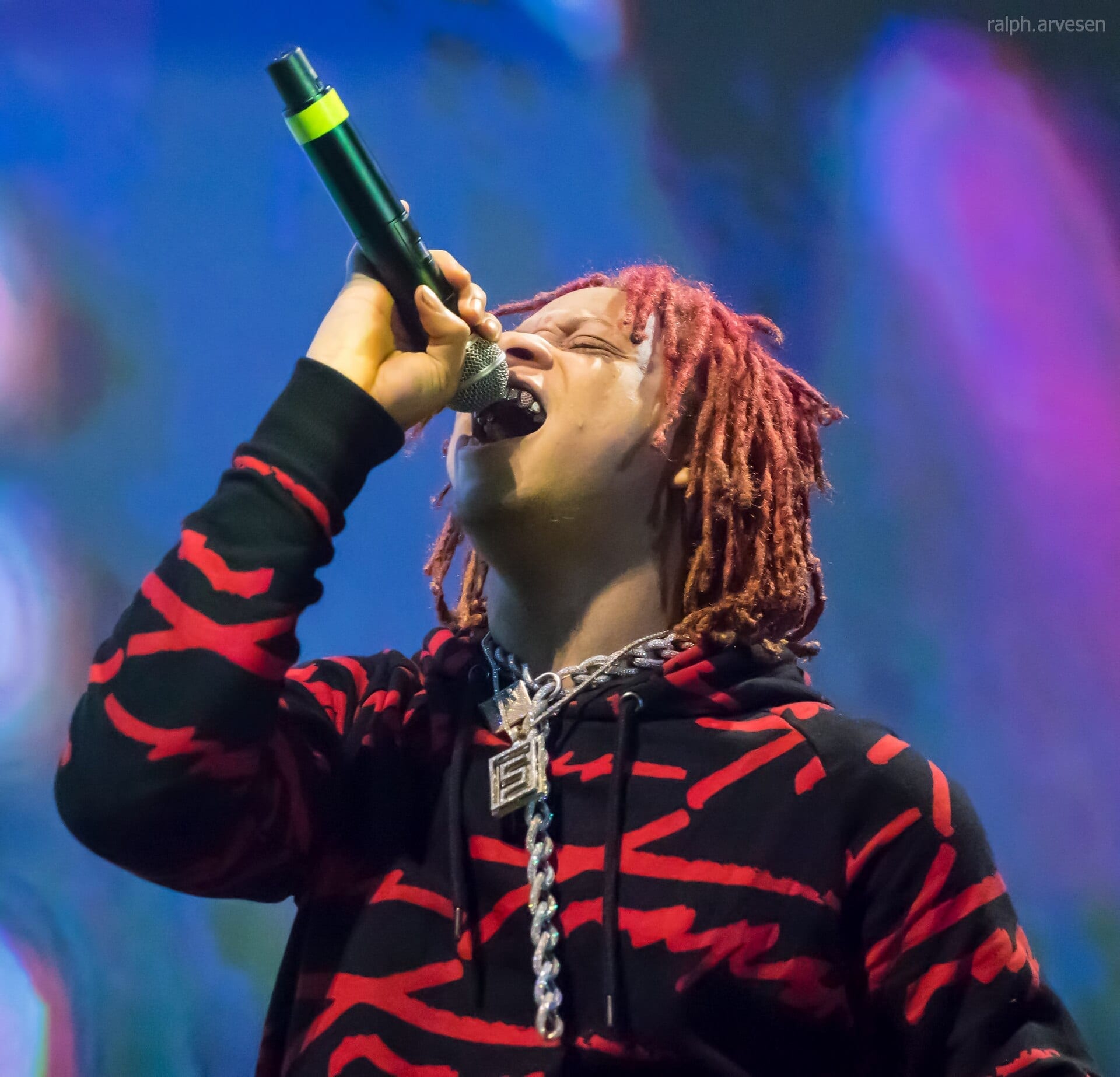 Trippie Redd, with red dreadlocks and a diamond chain, sings into a microphone held close to his mouth against a blue and purple stage-lit background.