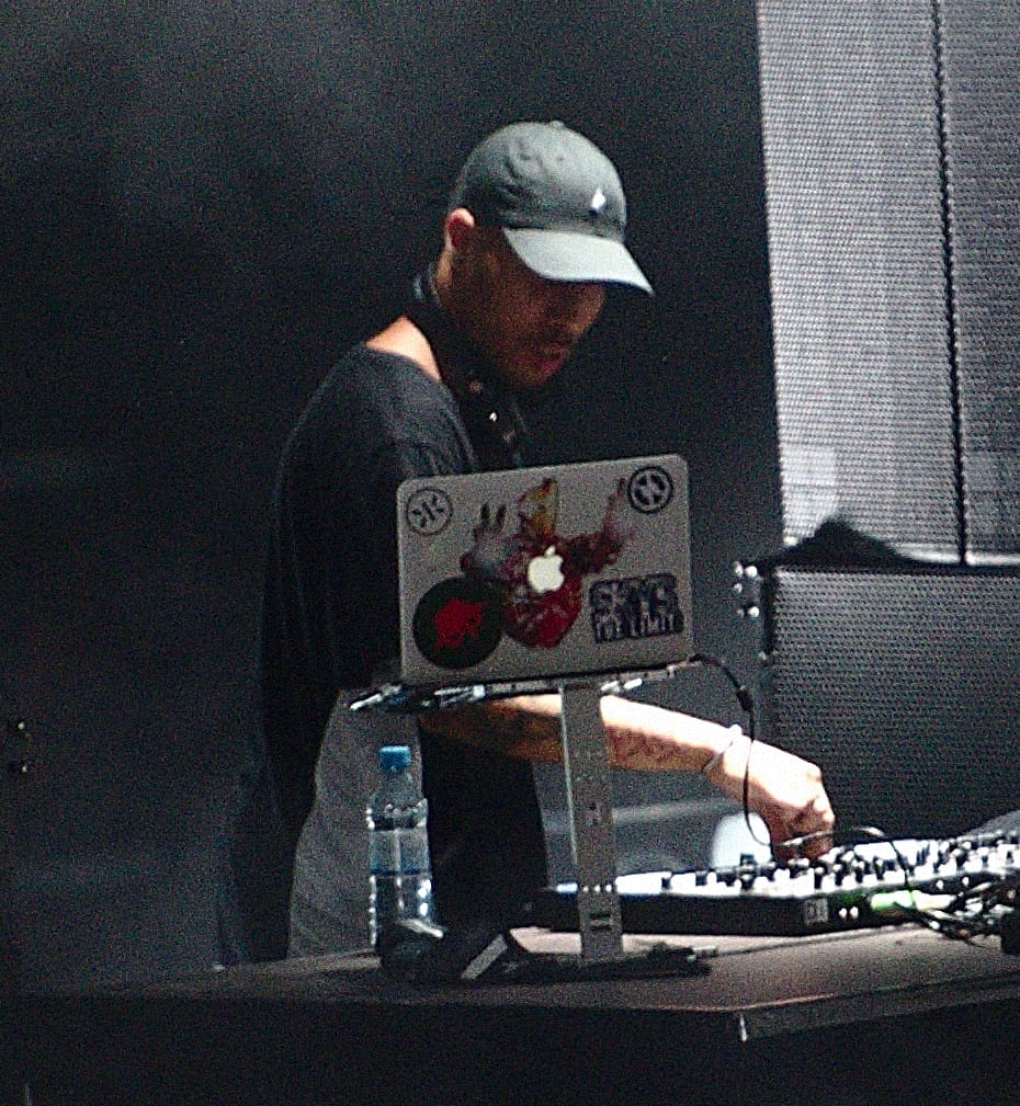 TroyBoi, wearing a cap and dark shirt, looks down while adjusting a mixer on a DJ table with a laptop propped in front of him.