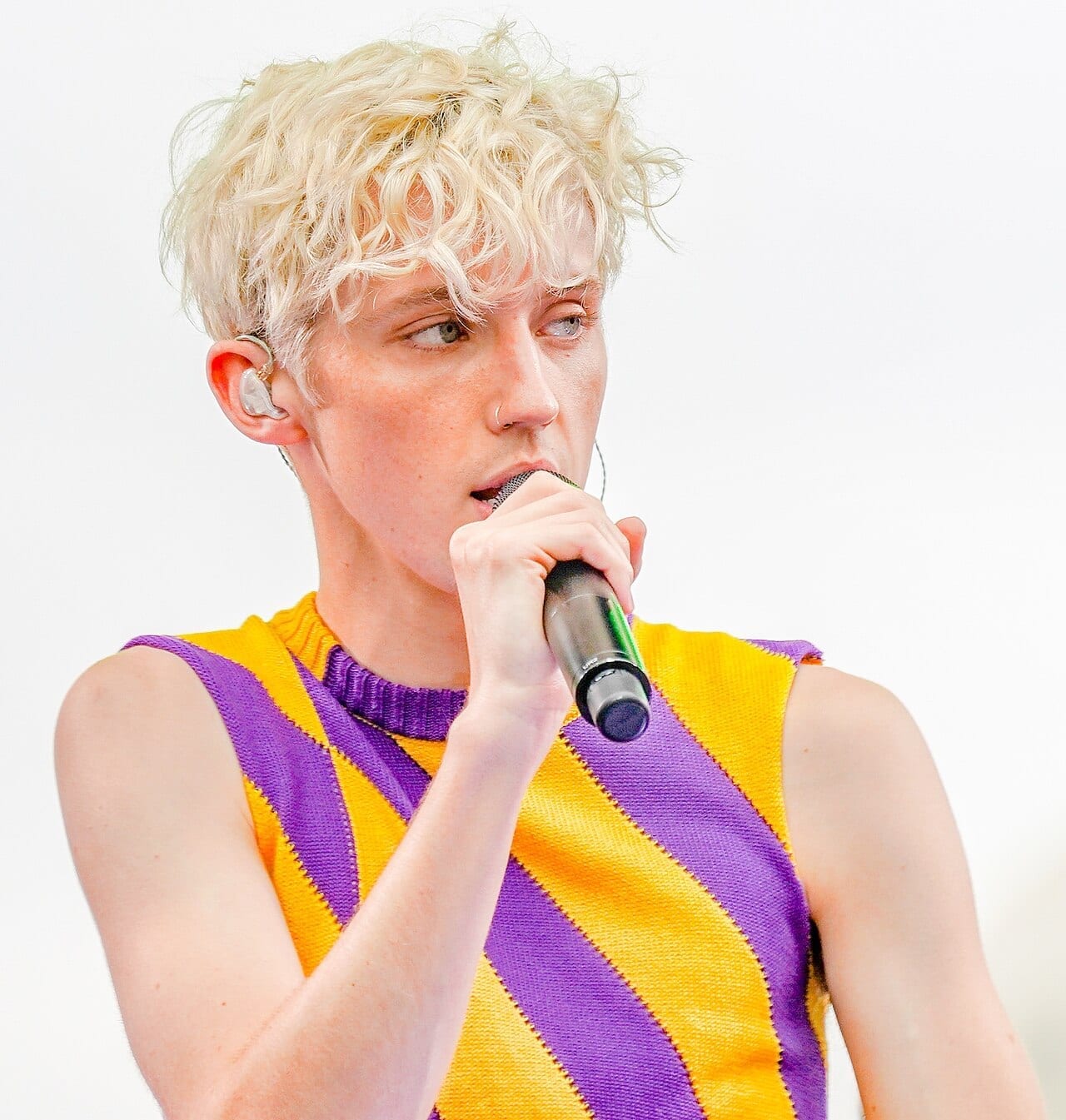 Troye Sivan sings into a handheld microphone against a white background, wearing a purple and yellow striped tank top.