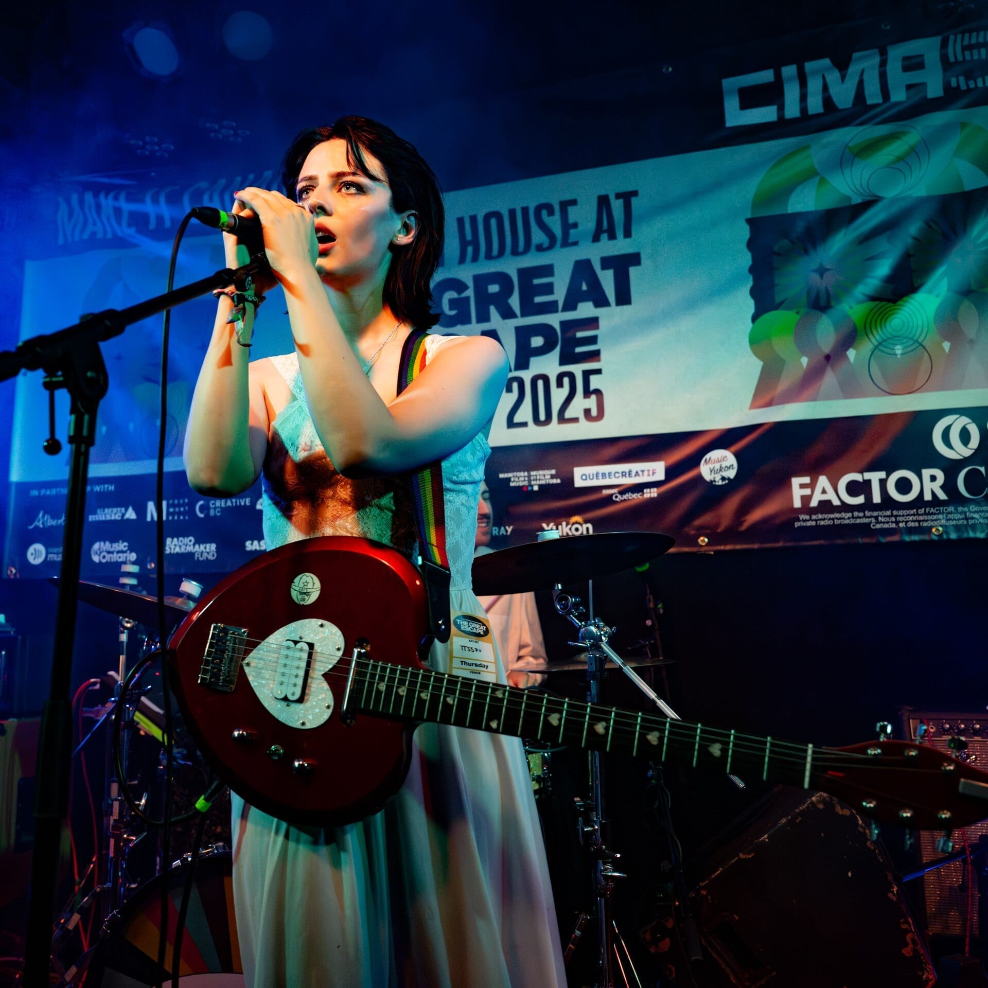 TTSSFU sings into a microphone while playing a red electric guitar, wearing a pale blue dress and an artist lanyard reading TTSSFU, with festival signage visible behind her.