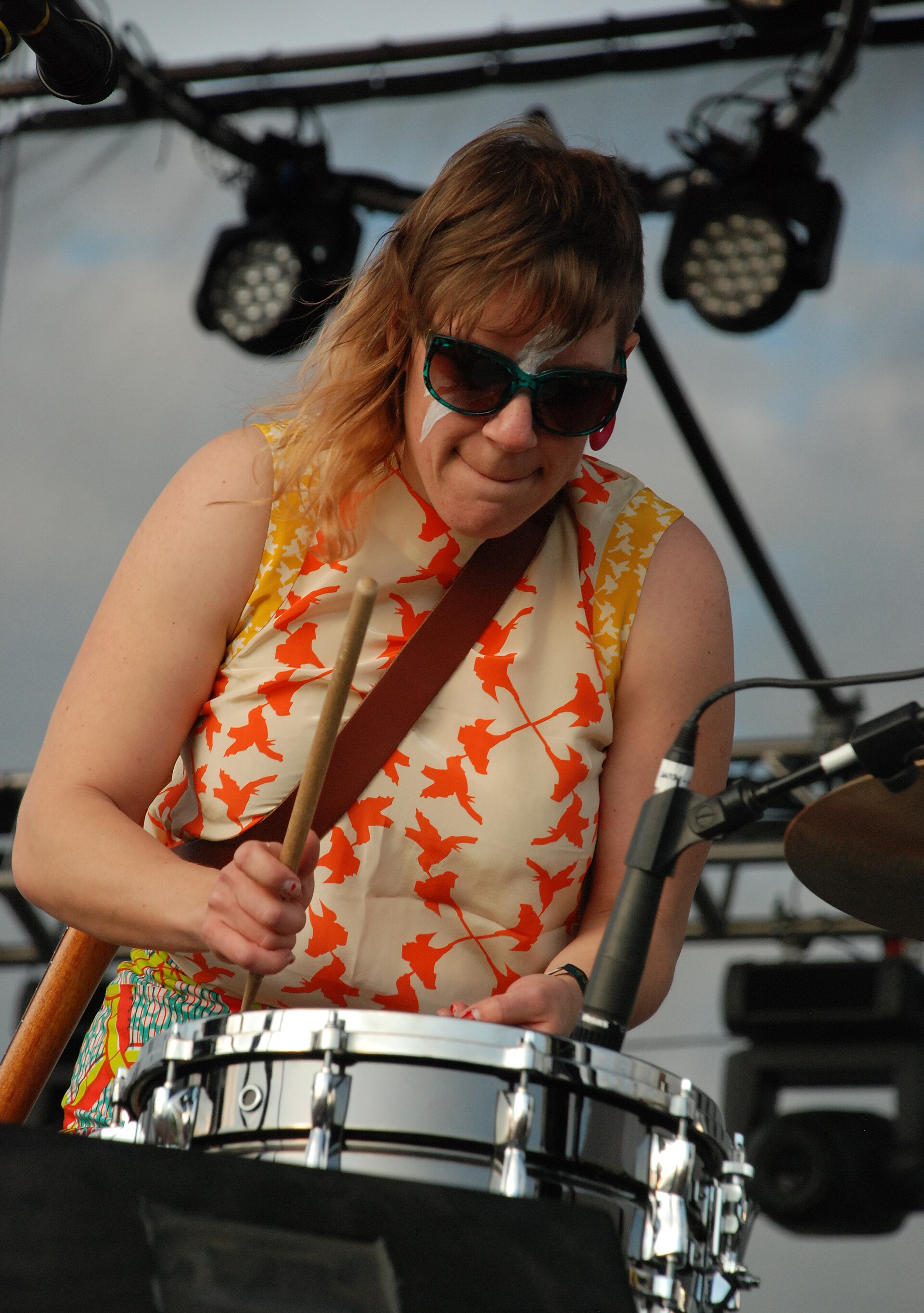 A drummer in sunglasses, face paint and a bird-print top hits a snare with one stick on an outdoor festival stage