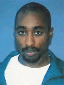 A close-up driver's license photograph of Tupac Shakur, a young Black man with a shaved head and thin mustache, wearing a blue jacket over a white shirt against a plain blue background.