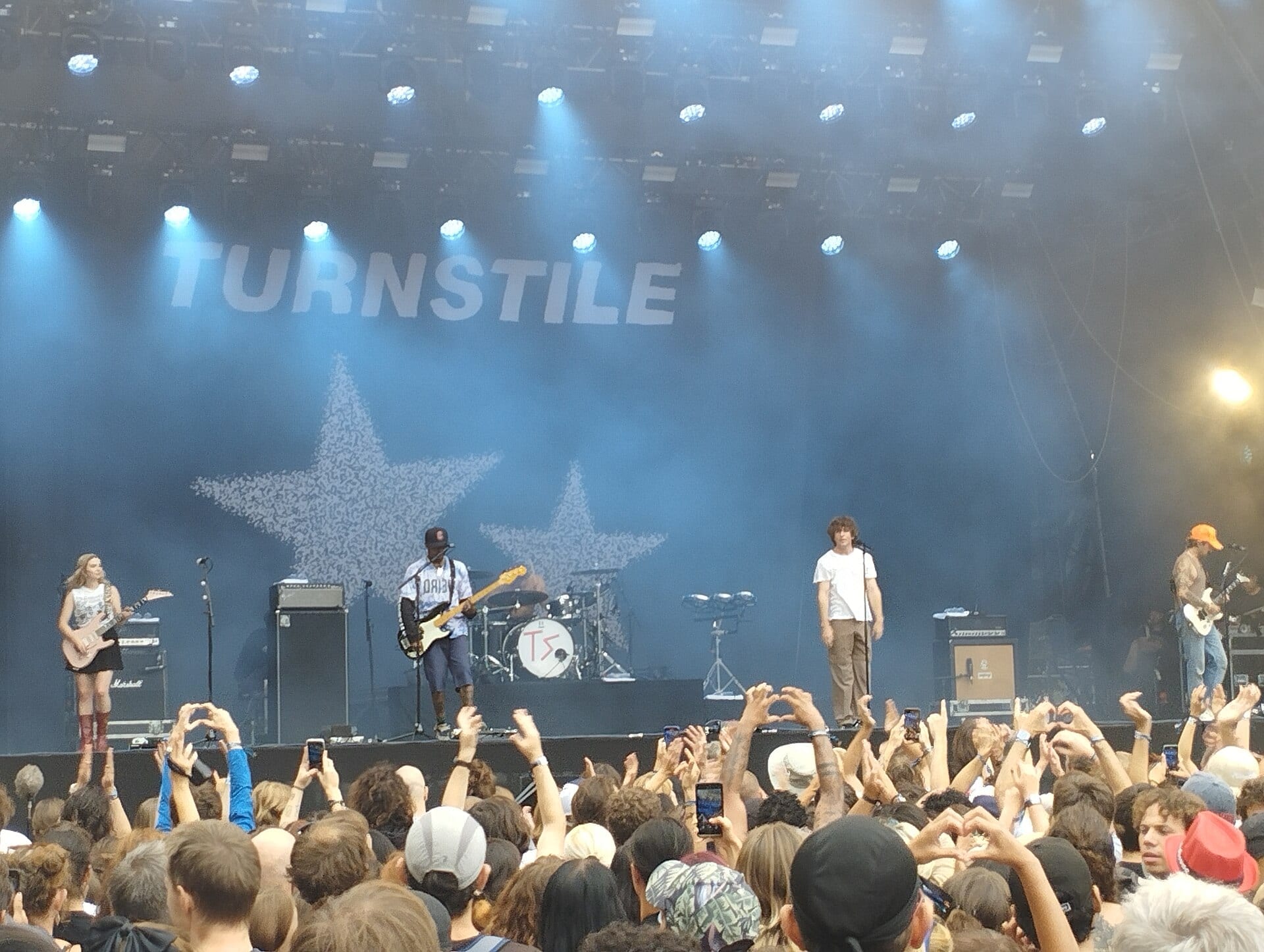 Turnstile play a festival stage under a large TURNSTILE banner and two star graphics, with fans holding up phones and making heart shapes in the crowd.