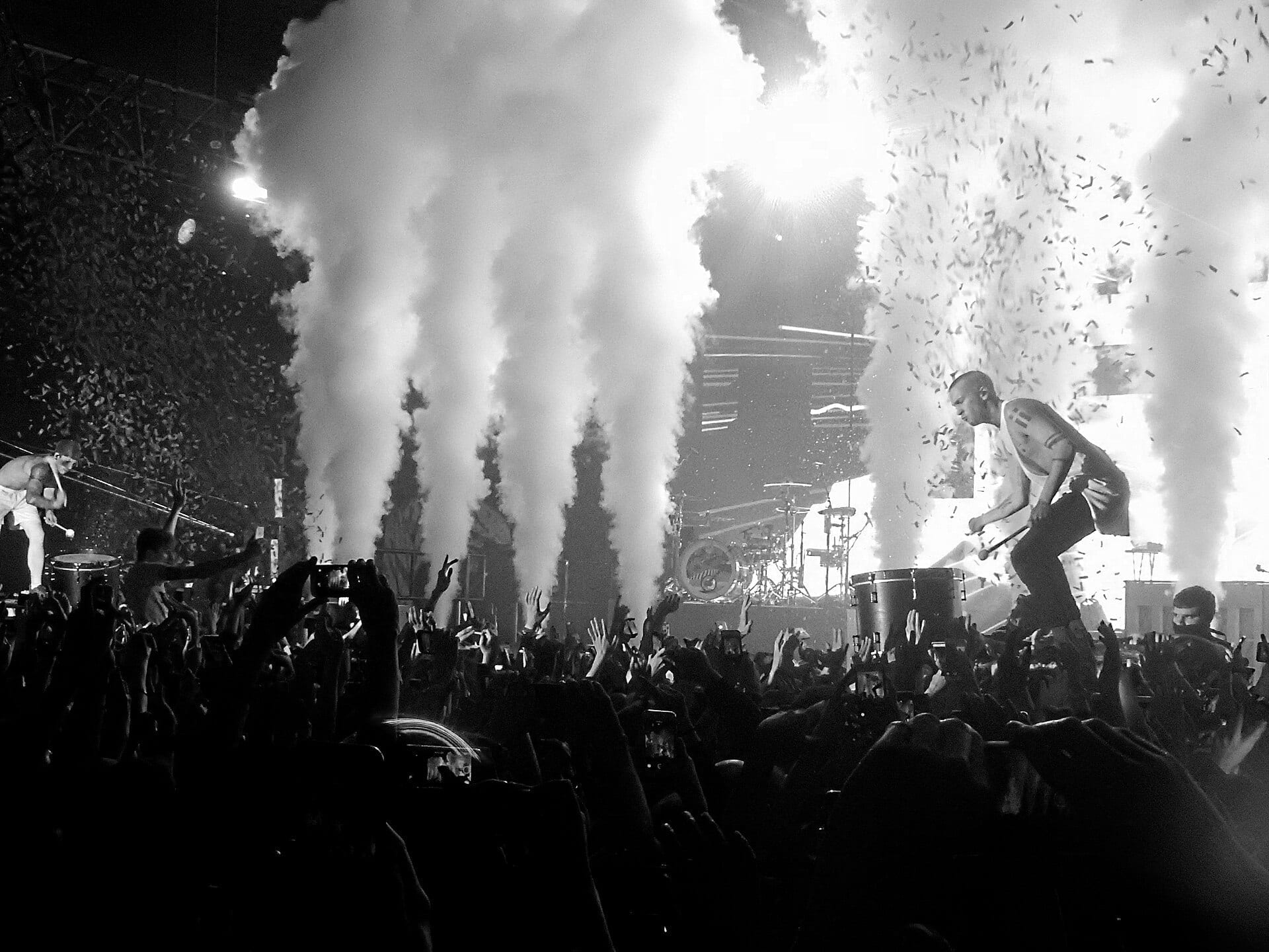 Twenty One Pilots perform on stage in black and white, engulfed in smoke and falling confetti as a shirtless band member with painted arms leaps near the drum riser.