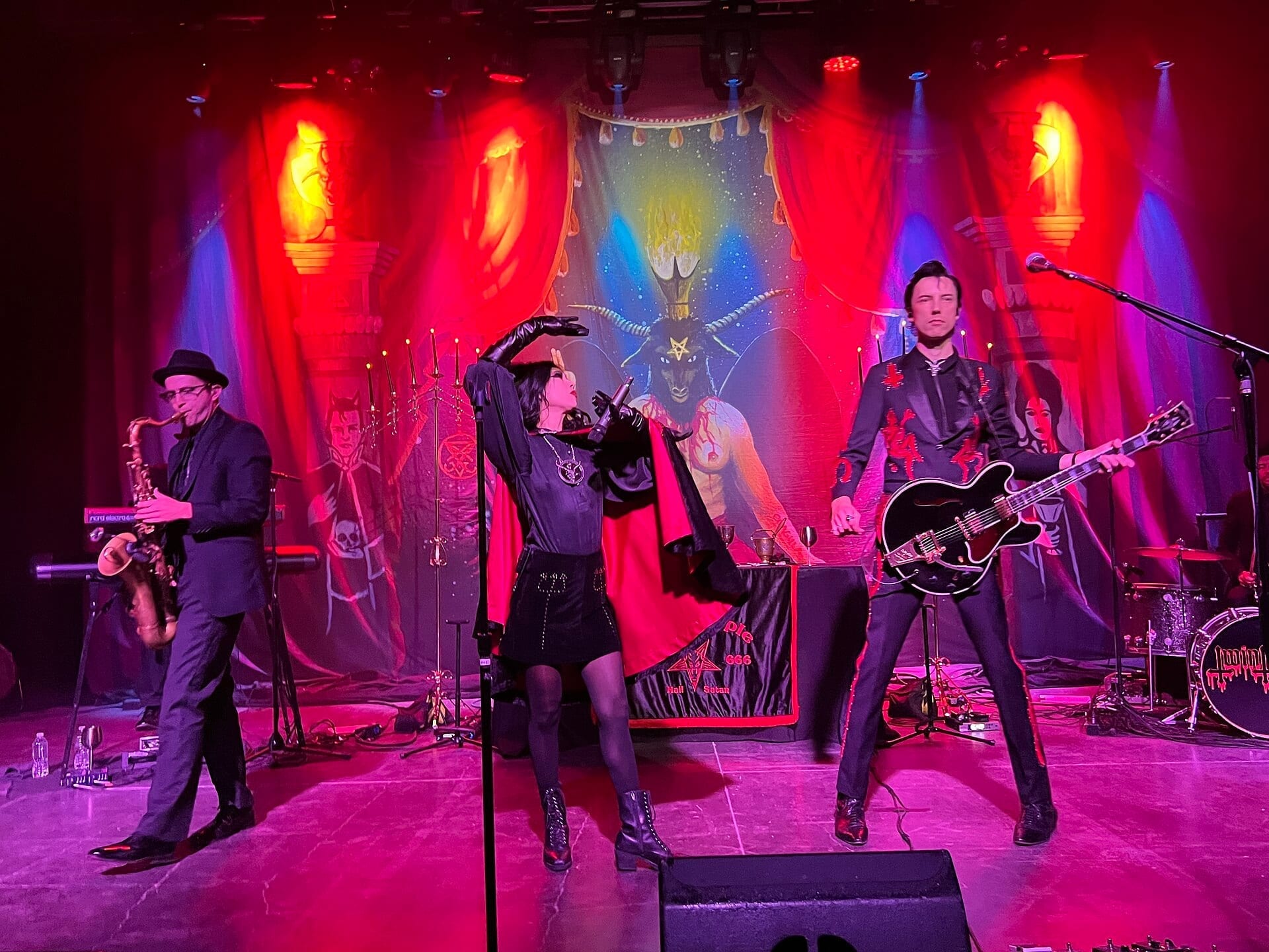 Twin Temple perform on a red-lit stage, the singer in a black cape reaching toward the microphone stand and a guitarist beside her, in front of a painted backdrop of a horned occult figure.