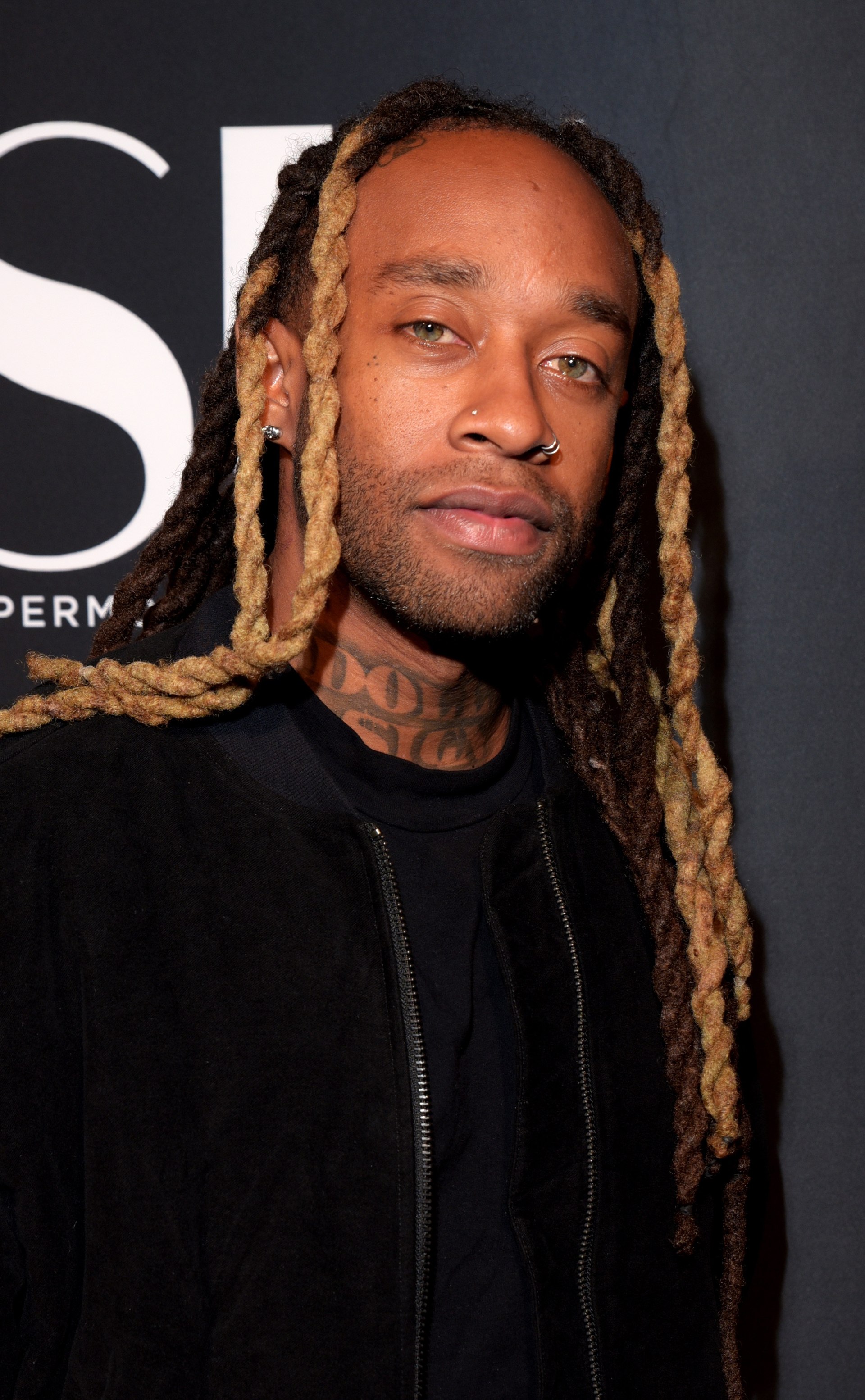 Ty Dolla $ign faces the camera in a black zip jacket, blonde-tipped locs falling past his shoulders, neck tattoos visible.