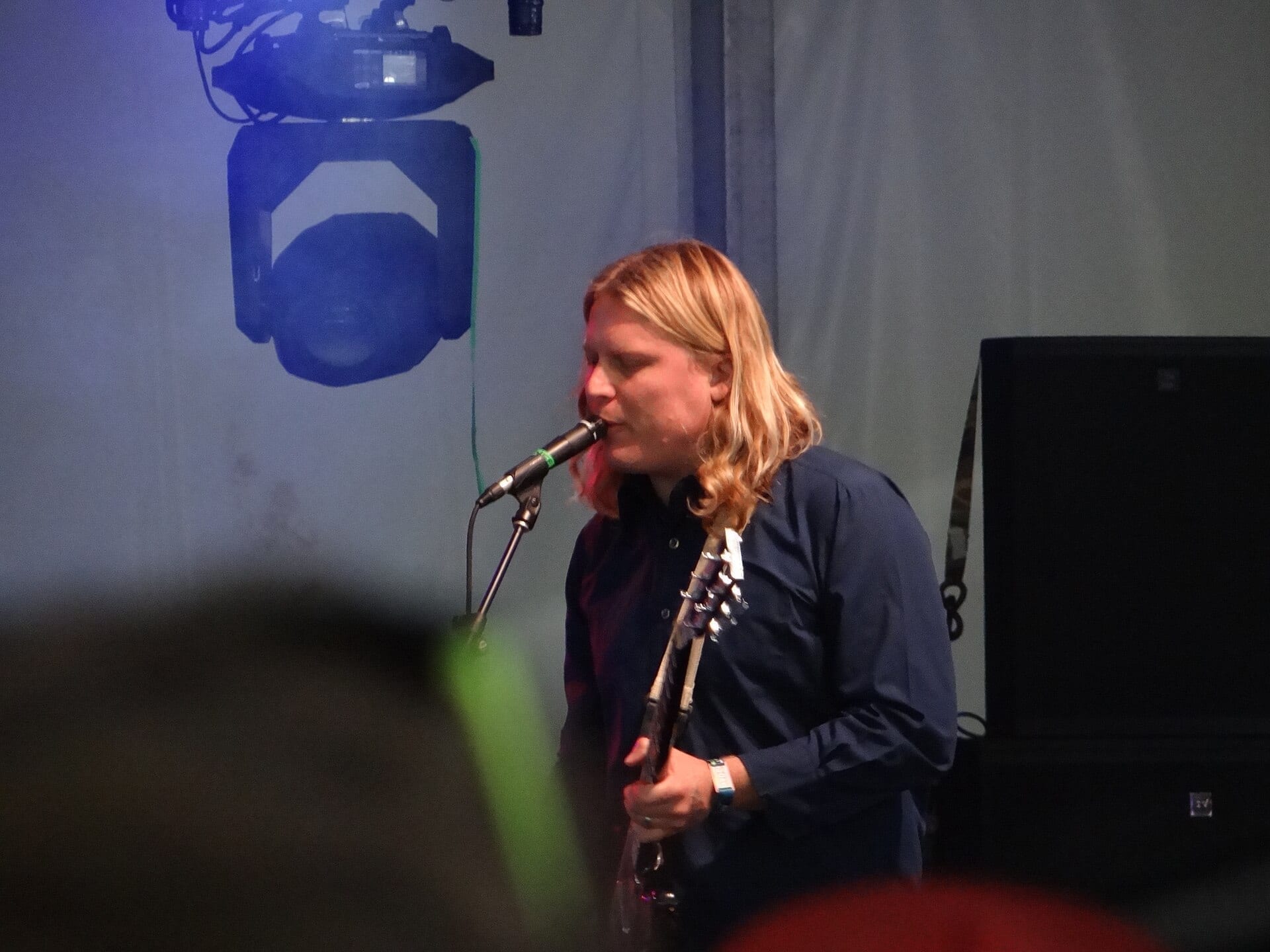 Ty Segall, with long blond hair, sings into a microphone while playing a guitar on a dim stage lit by a blue spotlight rig overhead.