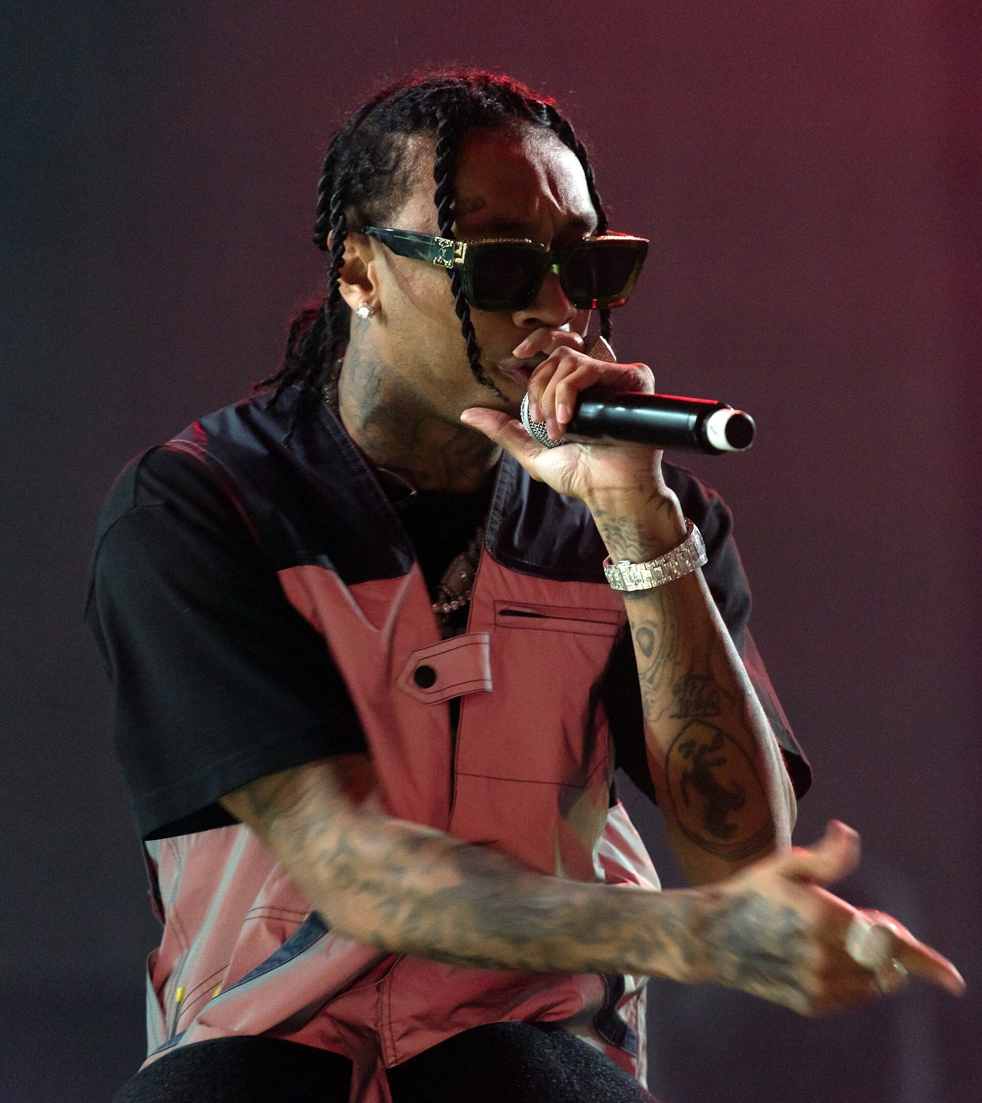 A close-up of Tyga performing into a microphone, wearing dark sunglasses and braids, arm tattoos visible, against a dark red background.