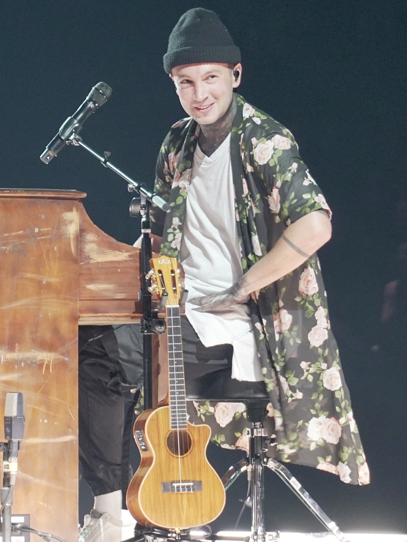 Tyler Joseph of Twenty One Pilots smiles in a beanie on a small satellite stage, a ukulele in front of him and heavy tattoos across his neck and arms.