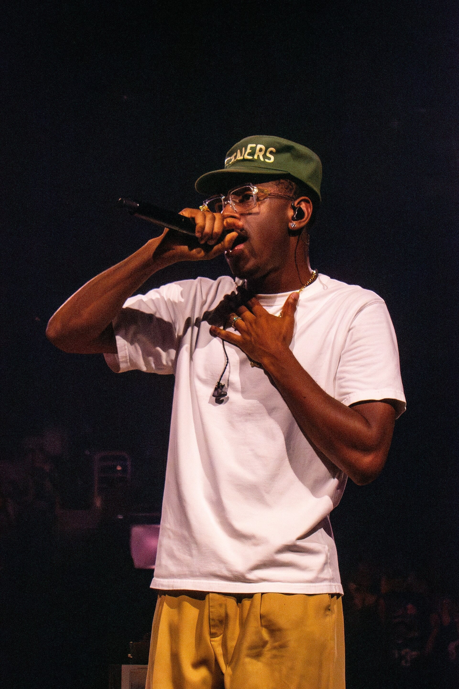 Tyler, The Creator raises a handheld microphone to his mouth mid-verse, one hand at his chest, wearing a green cap, clear-framed glasses, a white T-shirt and mustard trousers.