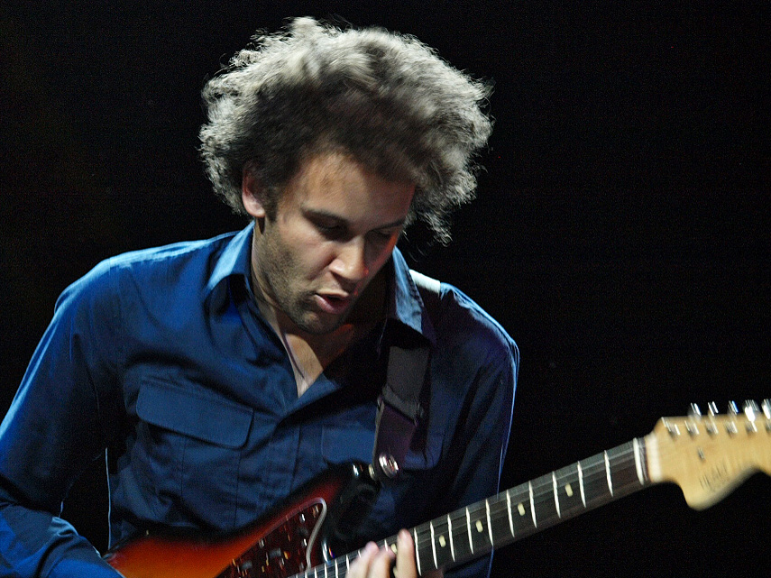 Tyondai Braxton looks down at the neck of a sunburst electric guitar, curly hair backlit, in a dark blue shirt against a black background.