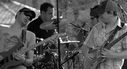 Umphrey's McGee performs outdoors, with two guitarists flanking a drummer, in a black and white photo.