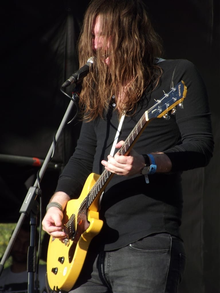 A long-haired Uncle Acid & The Deadbeats band member sings into a microphone while playing a yellow electric guitar on a shaded outdoor stage.